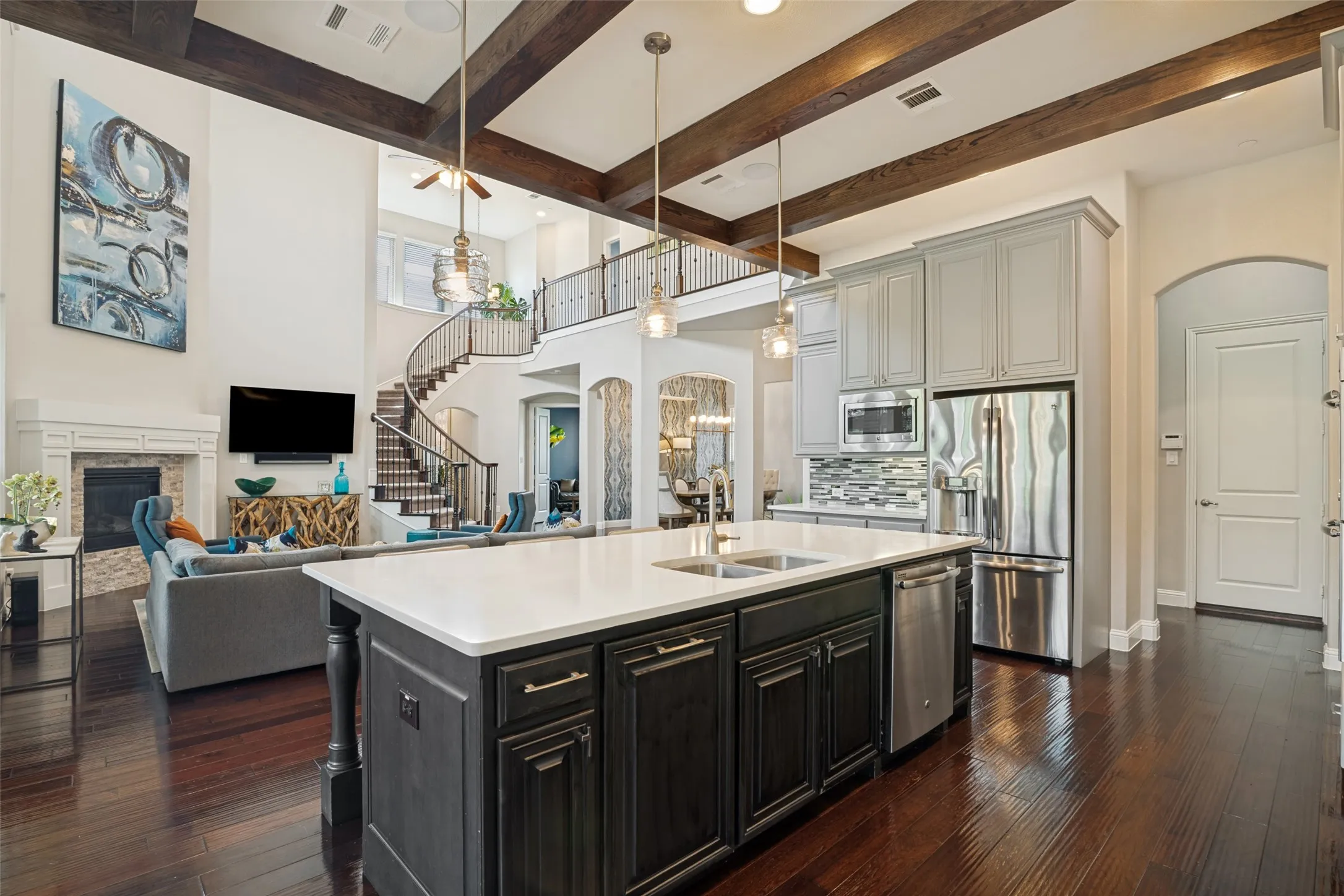 Gourmet kitchen with light gray cabinetry and stainless steel appliances, dark wood-finish flooring, white island countertop, and a two-story living area with a fireplace