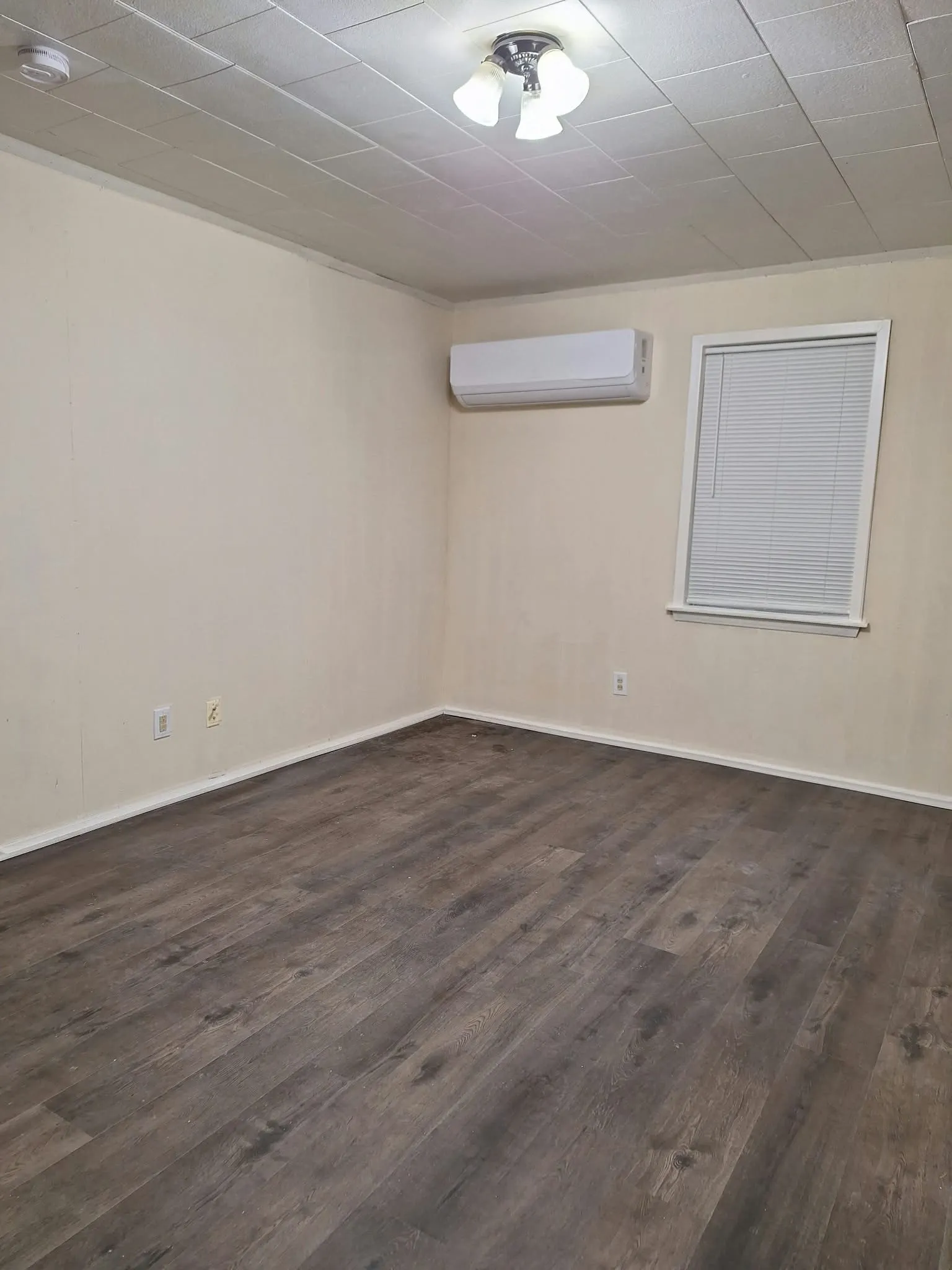 Wood-finish flooring, ceiling-mounted light fixture, window with blinds, wall-mounted climate control unit, and ceiling with acoustic tiles