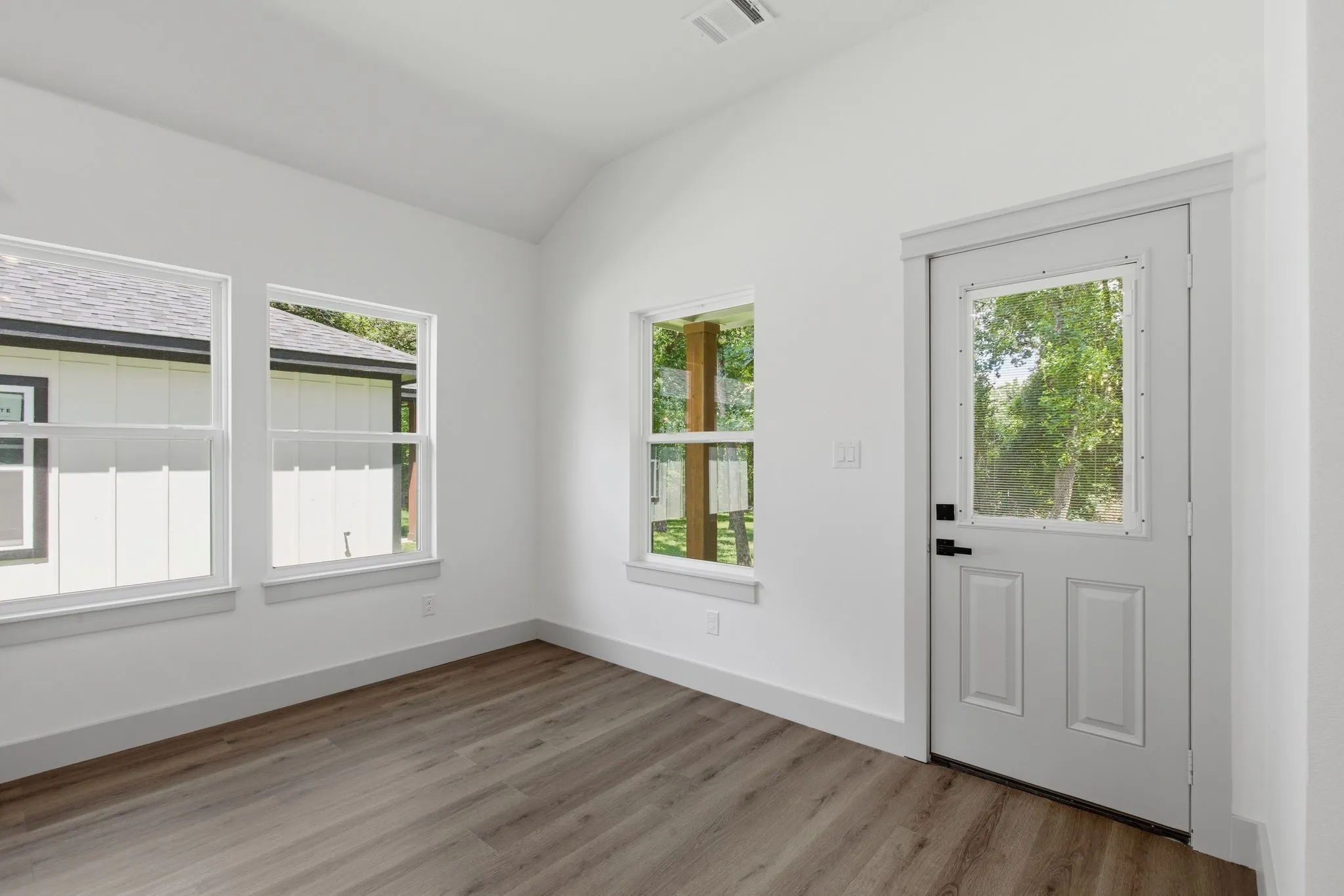 Vaulted ceiling, wood-finish flooring, multiple windows, white paneled door with glass insert, and white trim throughout
