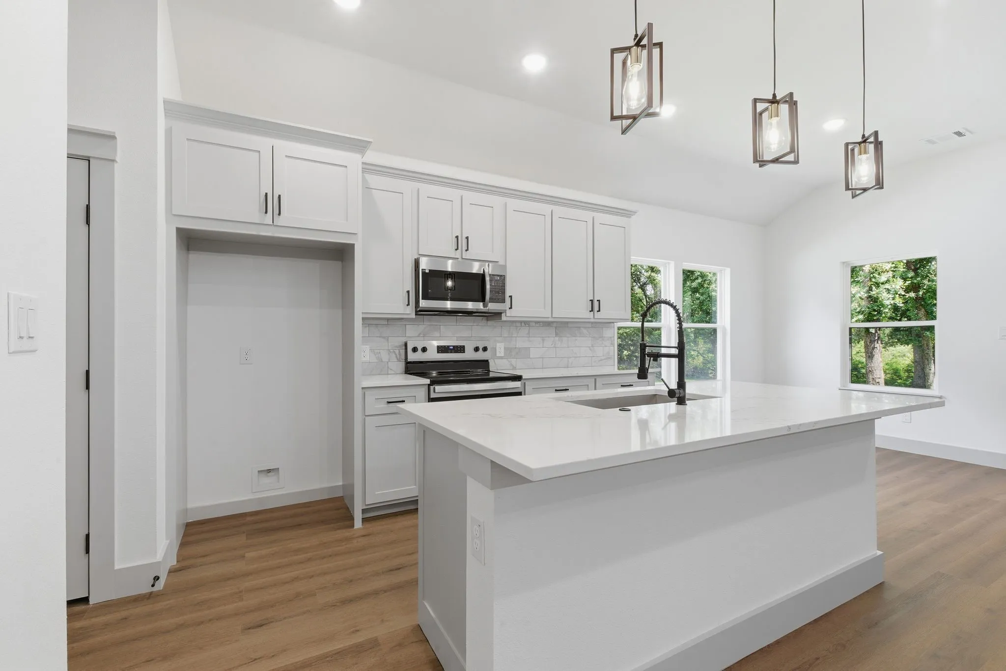 Kitchen featuring a large island with sink, stainless steel microwave, white shaker-style cabinetry, wood-finish flooring, and vaulted ceiling