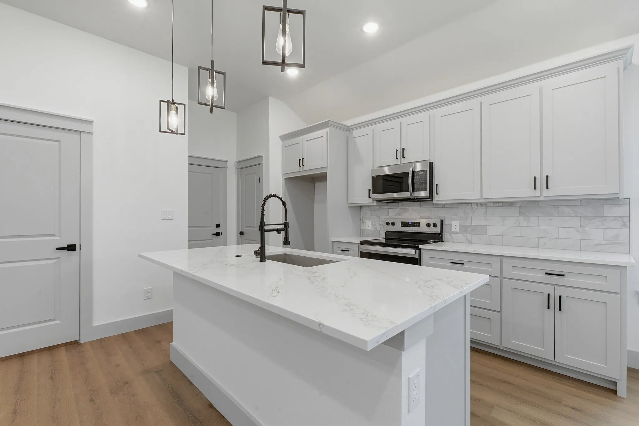 Kitchen featuring an island with a built-in sink, white countertops, light gray cabinetry with black hardware, a stainless steel microwave, and wood-finish flooring