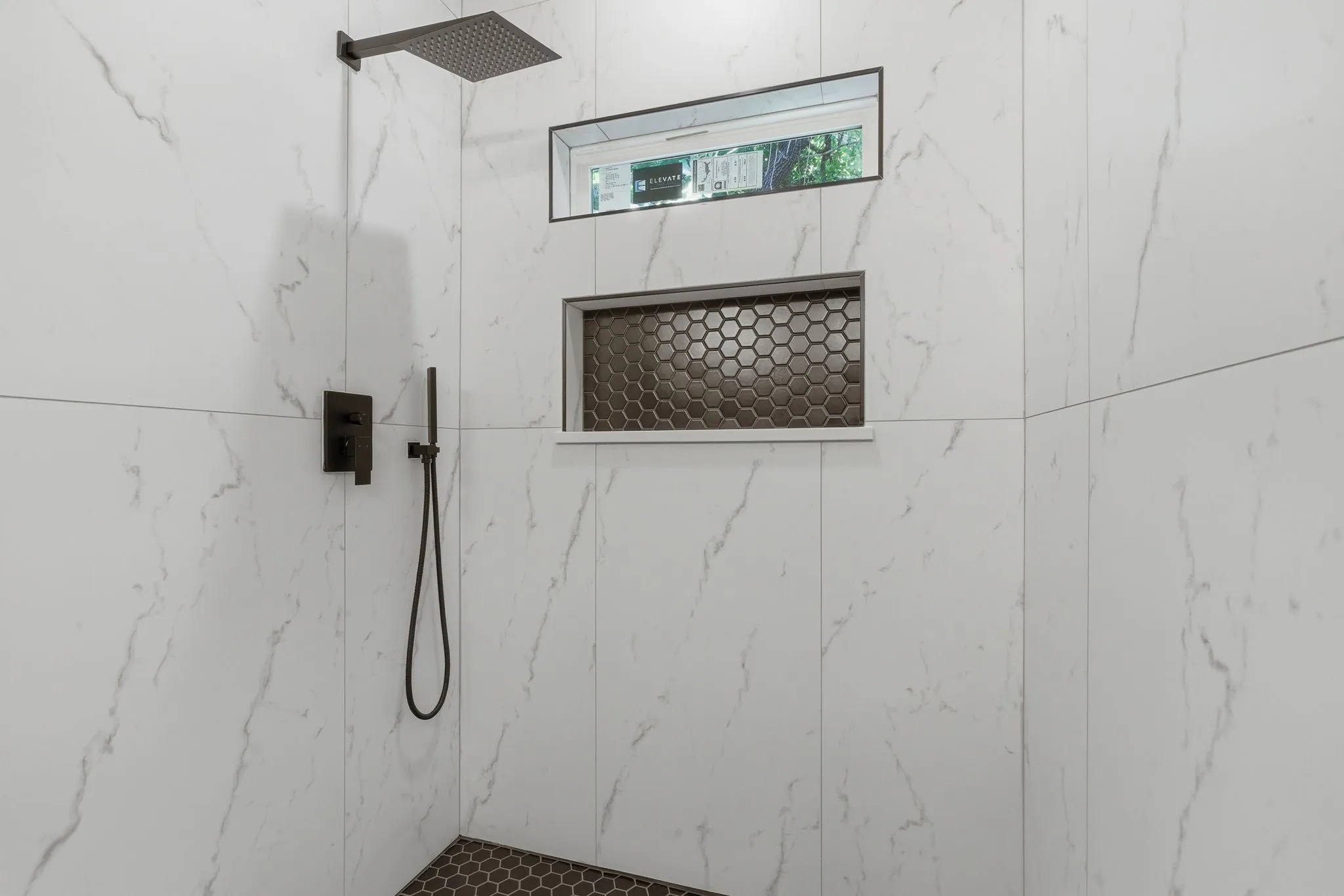 Shower featuring large format white tile walls, a matte black rain shower head, a matte black handheld shower, a built-in shower niche with hexagonal tile, and a privacy window