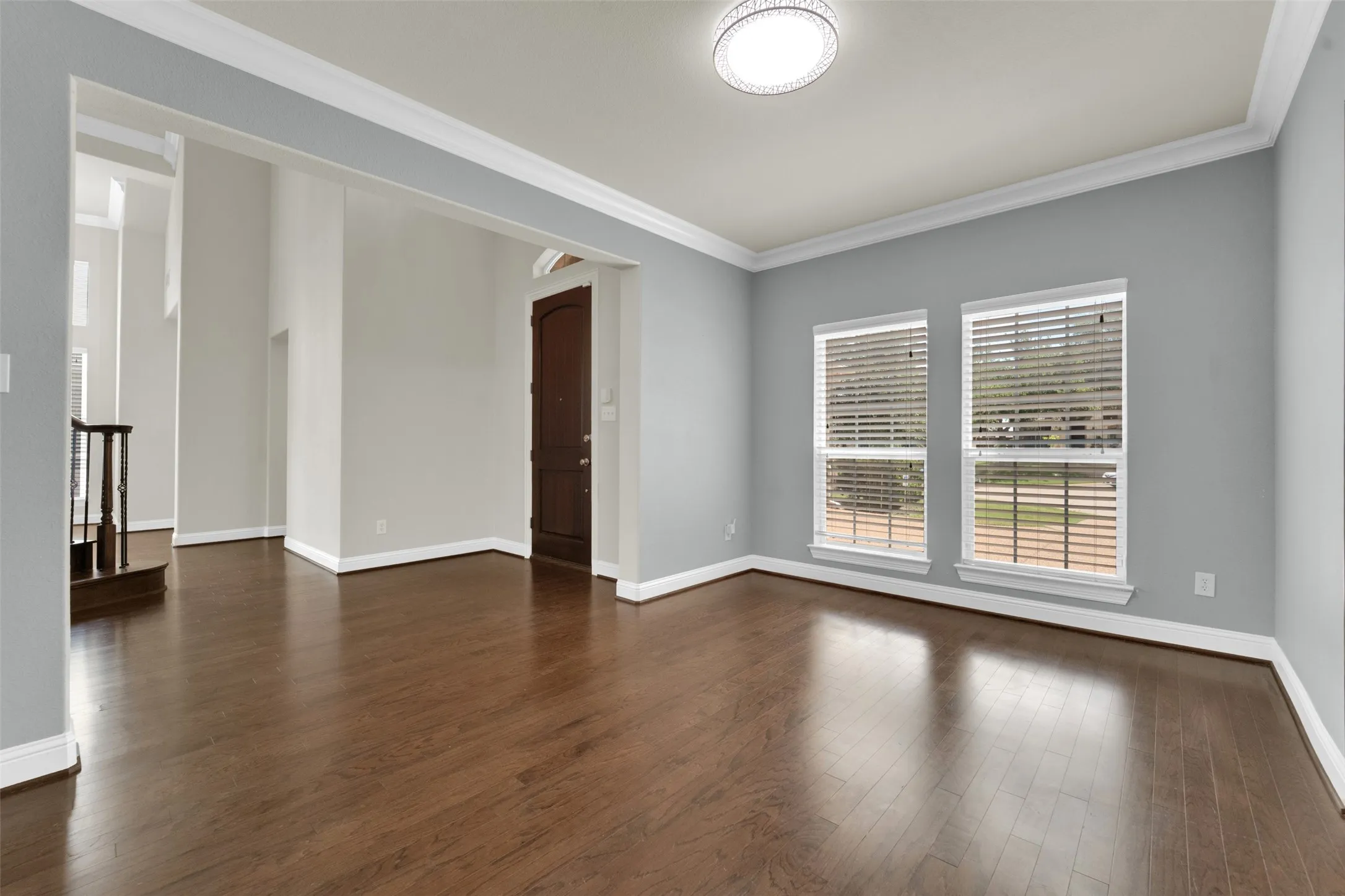 Dark wood-finish flooring, two large windows with blinds, crown molding, and a decorative flush-mount ceiling light