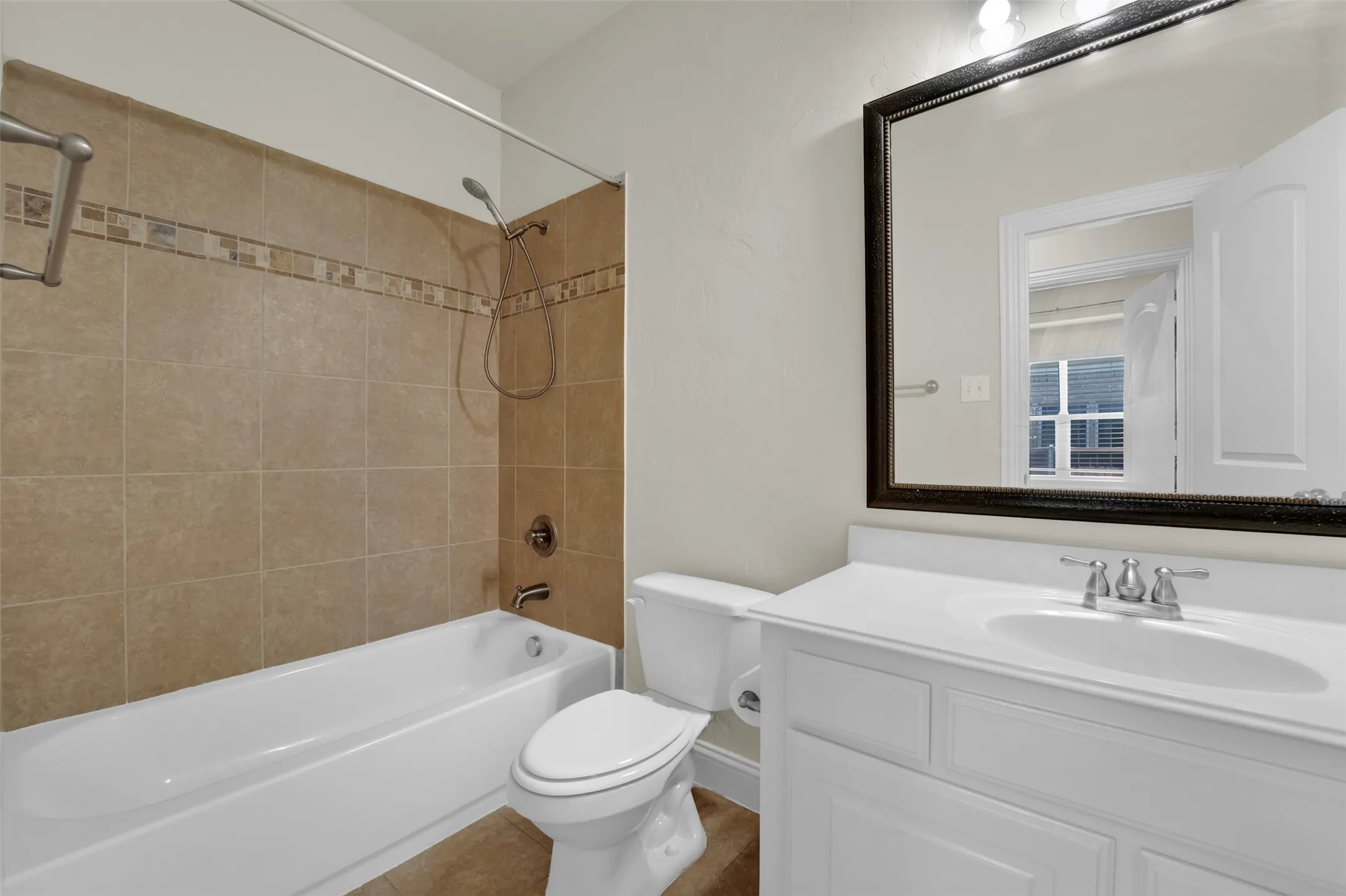 Tiled shower surround with decorative accent band, white bathtub, vanity with integrated sink, chrome fixtures, and framed mirror