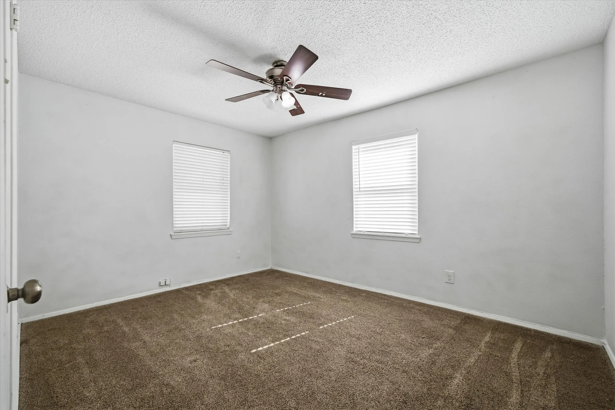 Carpeted flooring, two windows with blinds, ceiling fan with light fixture, and neutral wall paint