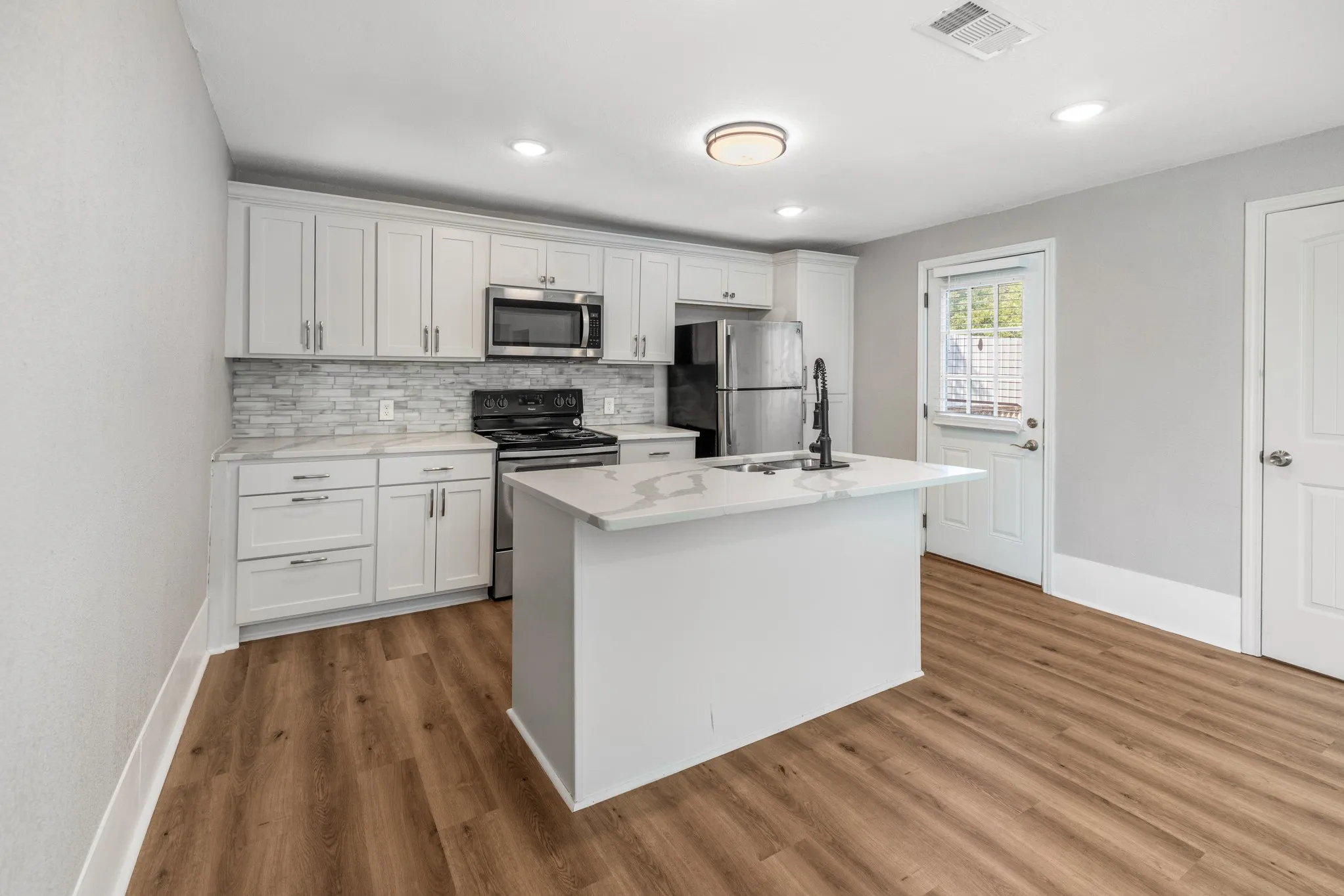 Kitchen featuring white shaker cabinetry, stainless steel appliances, a center island with sink, quartz countertops, and wood-finish flooring