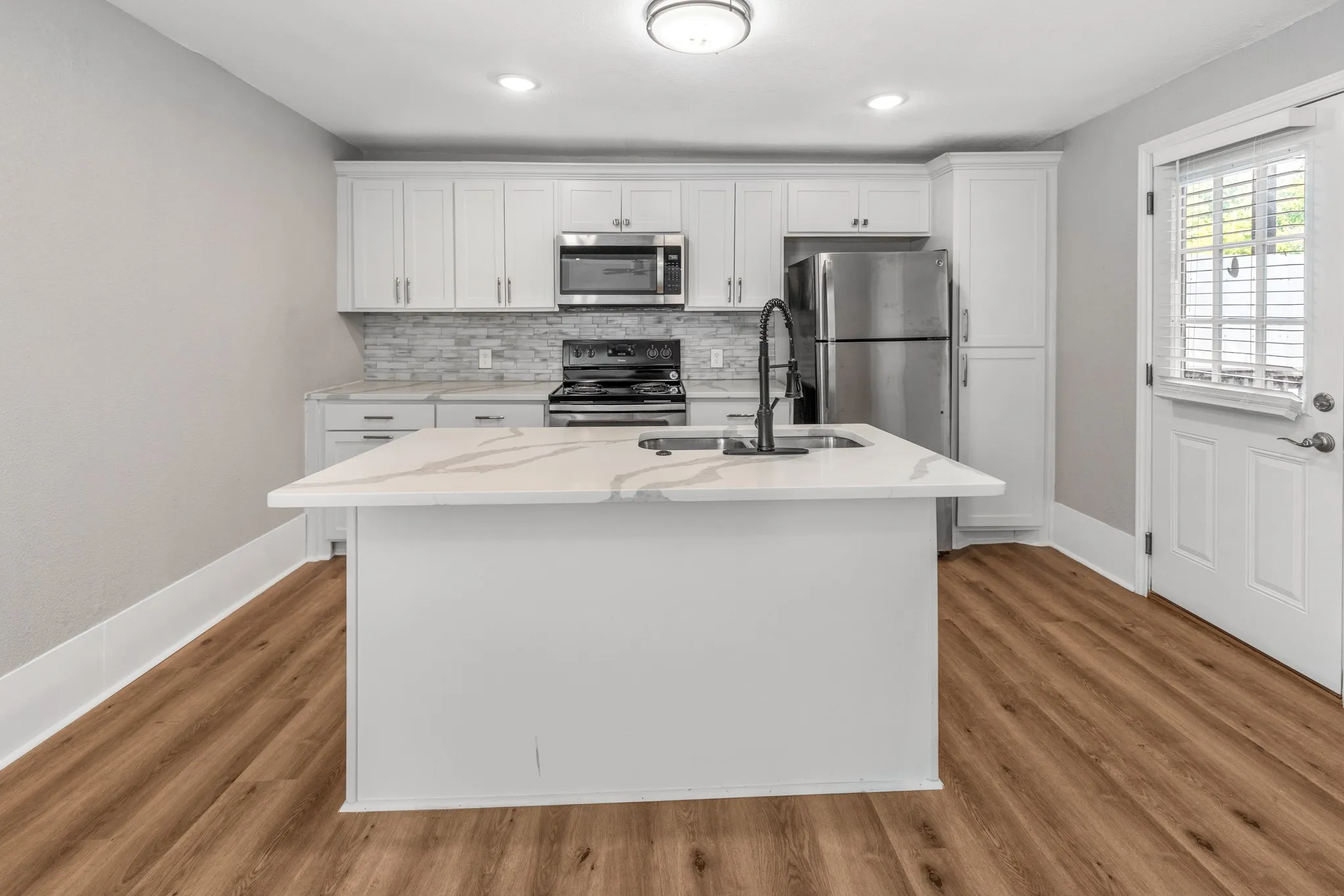 Kitchen featuring a central island with a sink, white cabinetry, stainless steel appliances, wood-finish flooring, and a paneled door