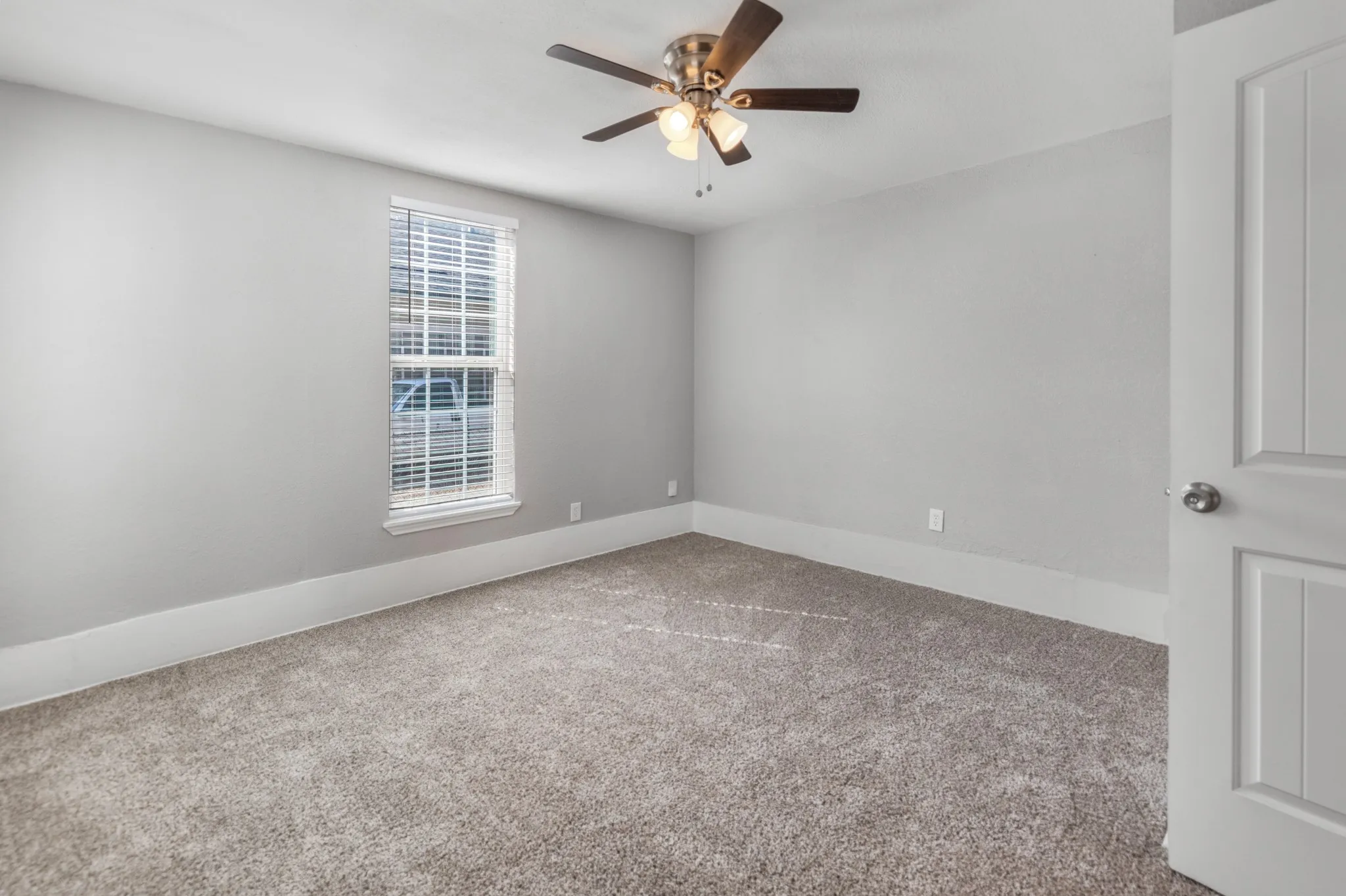 Neutral tone wall paint, textured carpet flooring, ceiling fan with integrated lighting, window with blinds, and a paneled door