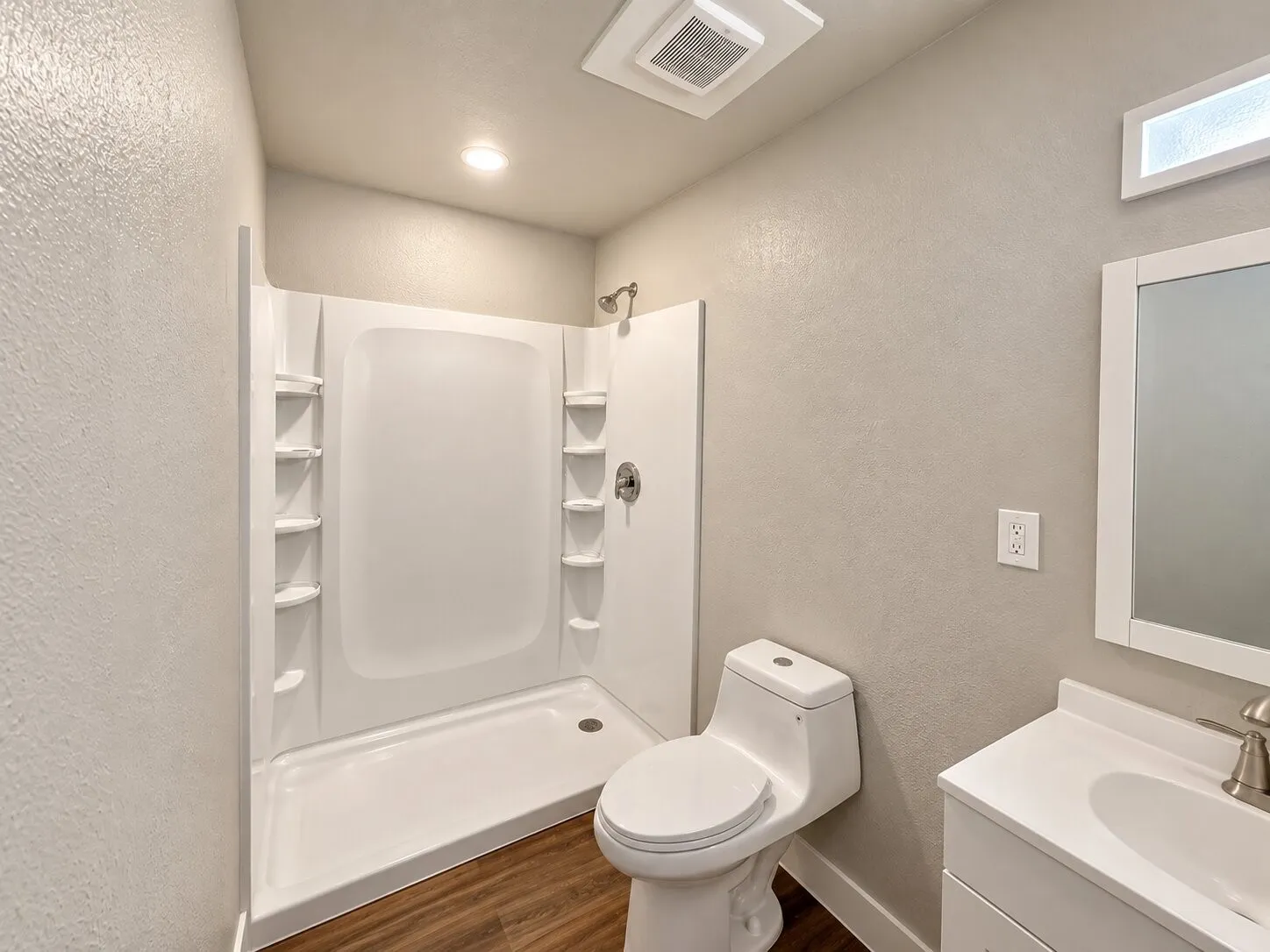 White shower stall with built-in shelving, wood-finish flooring, white toilet, single vanity with white countertop, and brushed nickel fixtures