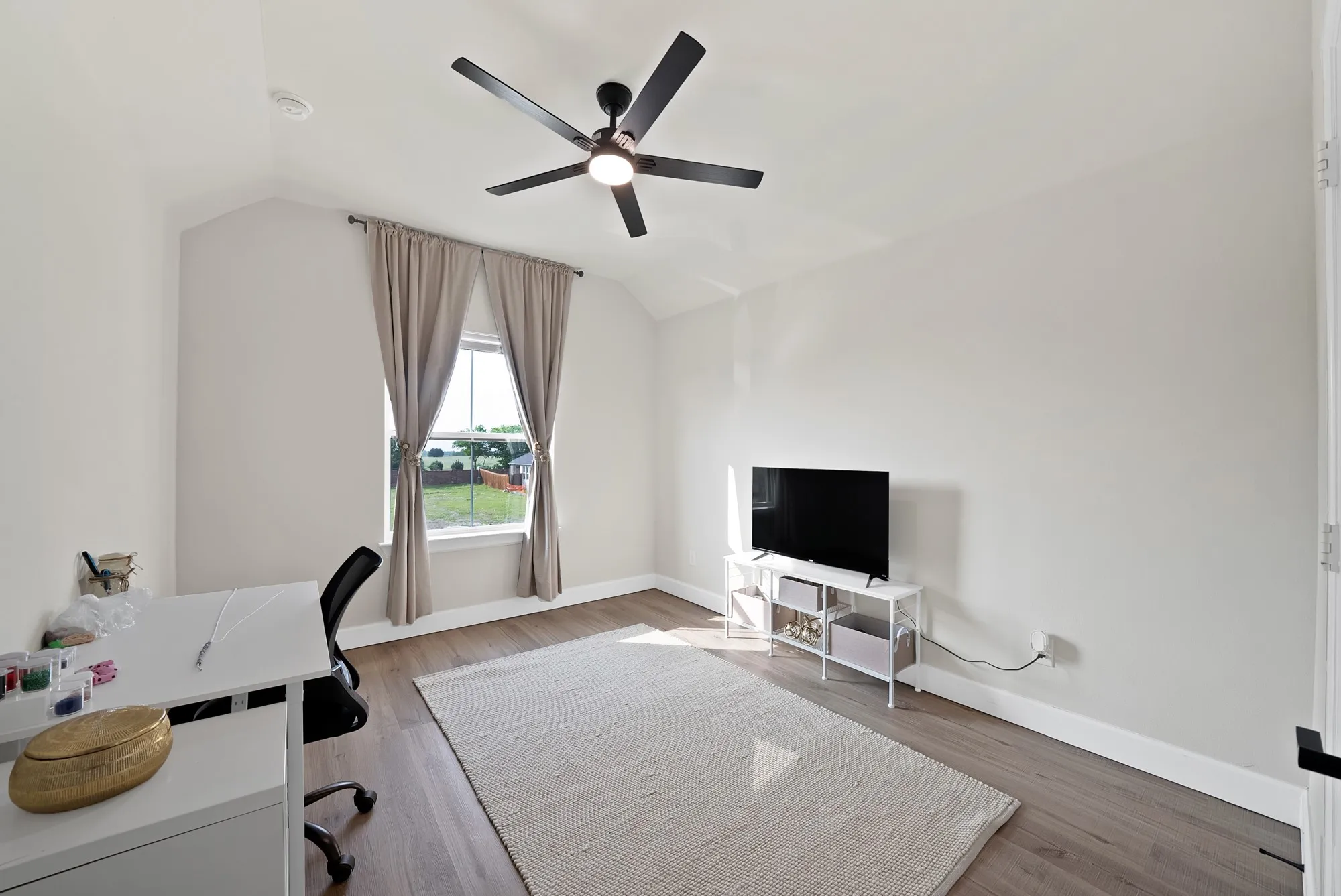 Vaulted ceiling with a black ceiling fan, wood-finish flooring, and a single window with curtains