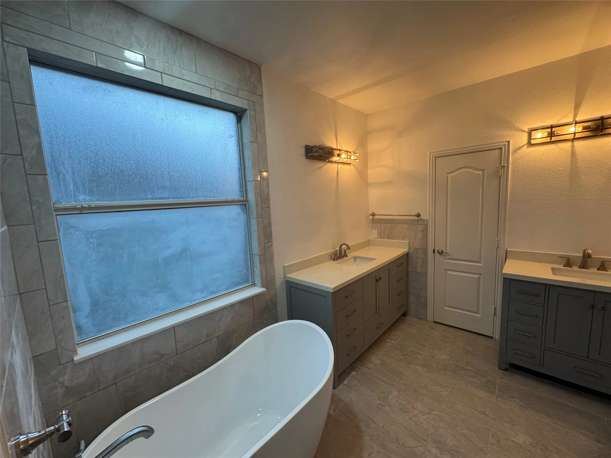 Porcelain tile flooring, freestanding soaking tub, dual vanity with light-toned countertops, and industrial-style wall sconces