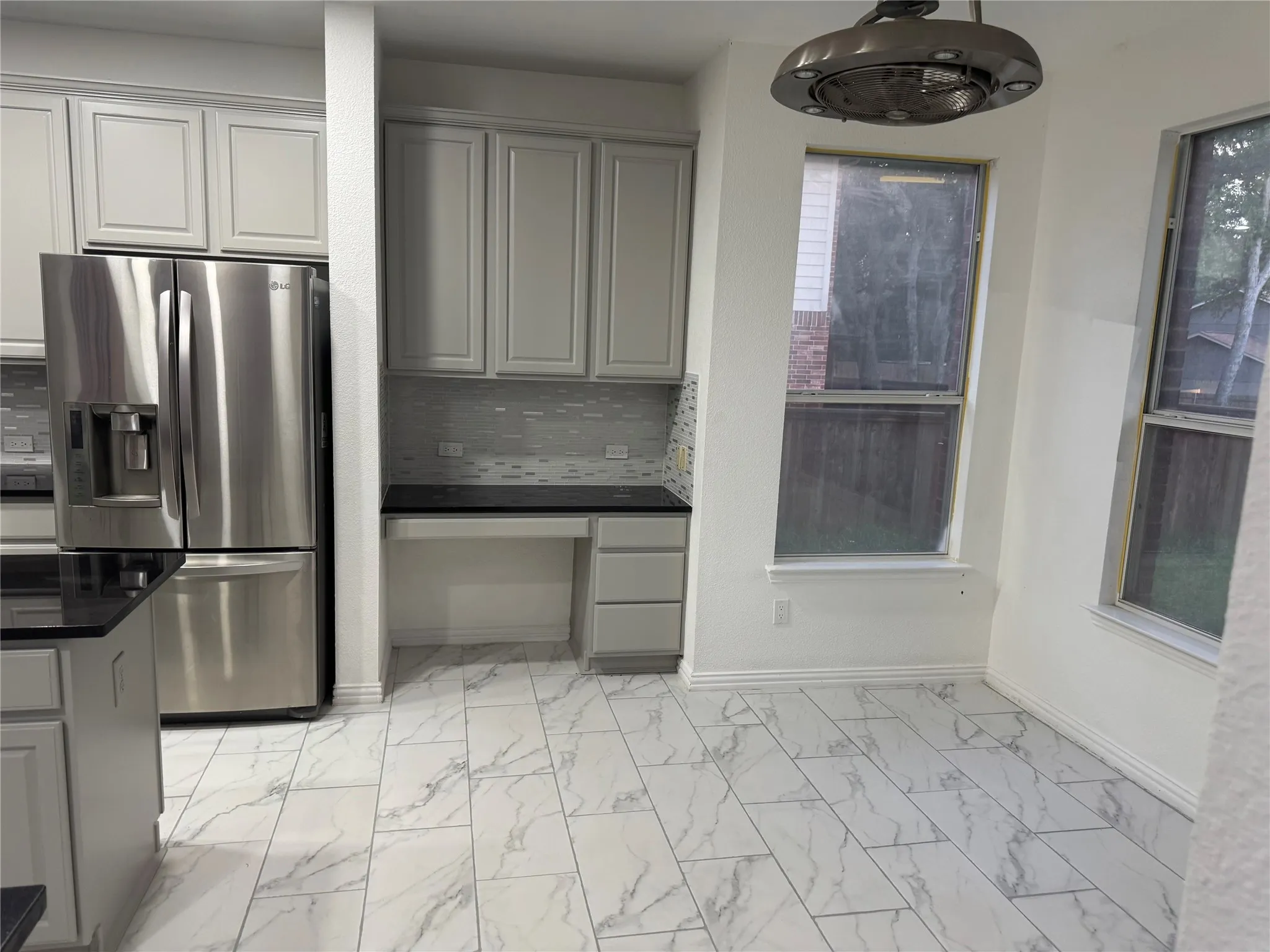 White and gray tile flooring, light gray cabinetry, stainless steel refrigerator, black countertops, and a built-in desk area