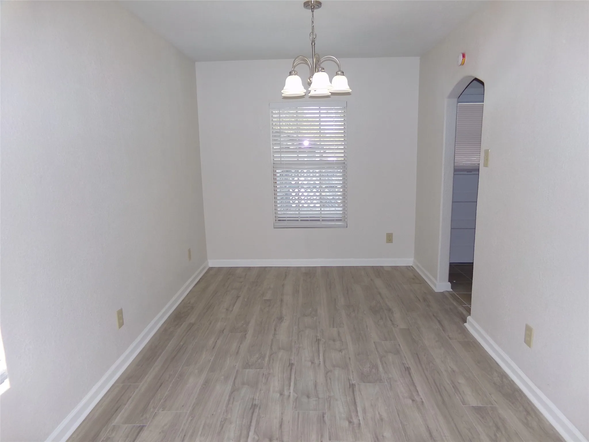 Wood-finish flooring, white baseboards, recessed window with blinds, arched doorway, and a flush-mount chandelier
