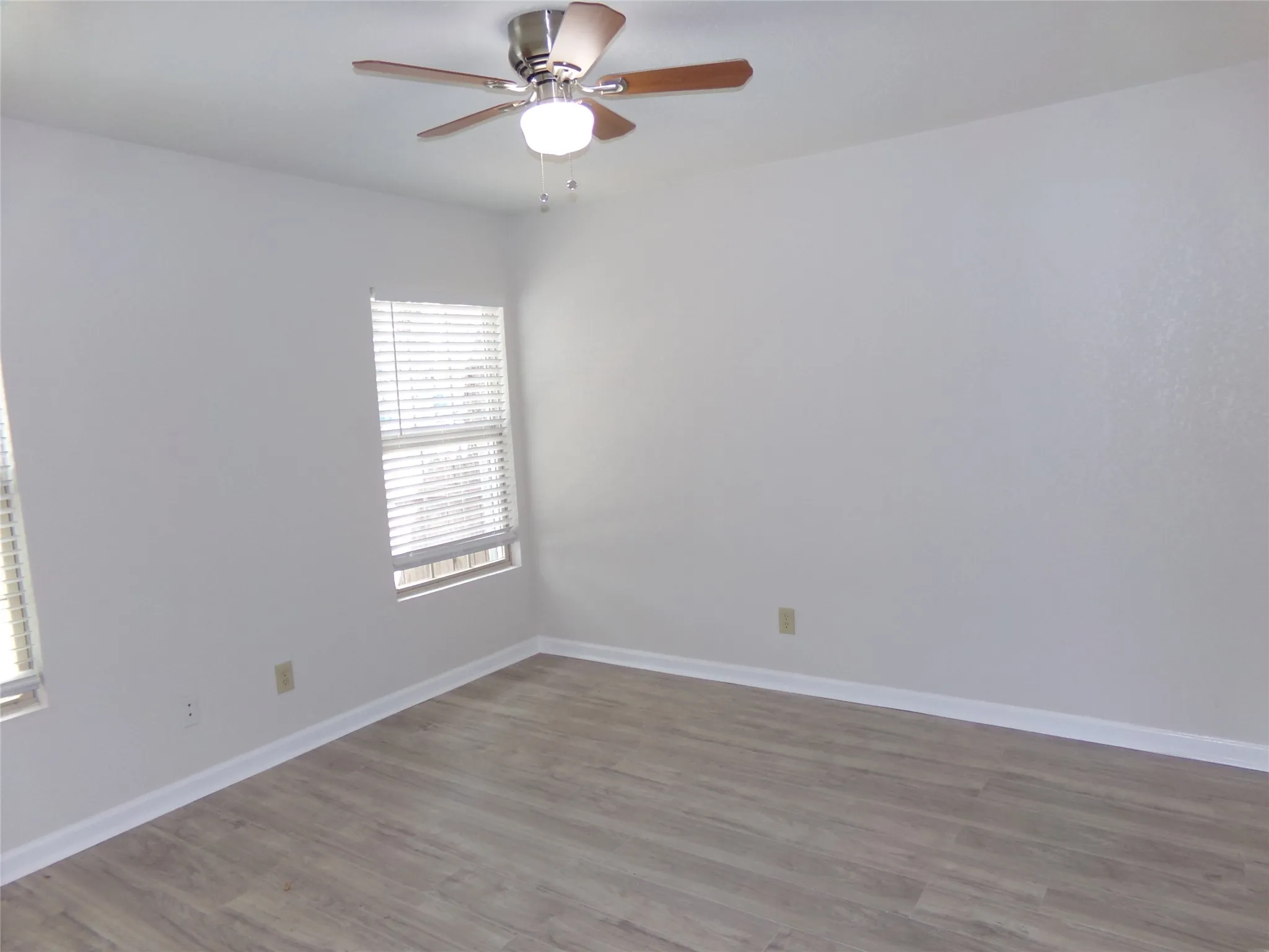 Light wood-finish flooring, white walls, two windows with blinds, and a ceiling fan with integrated lighting