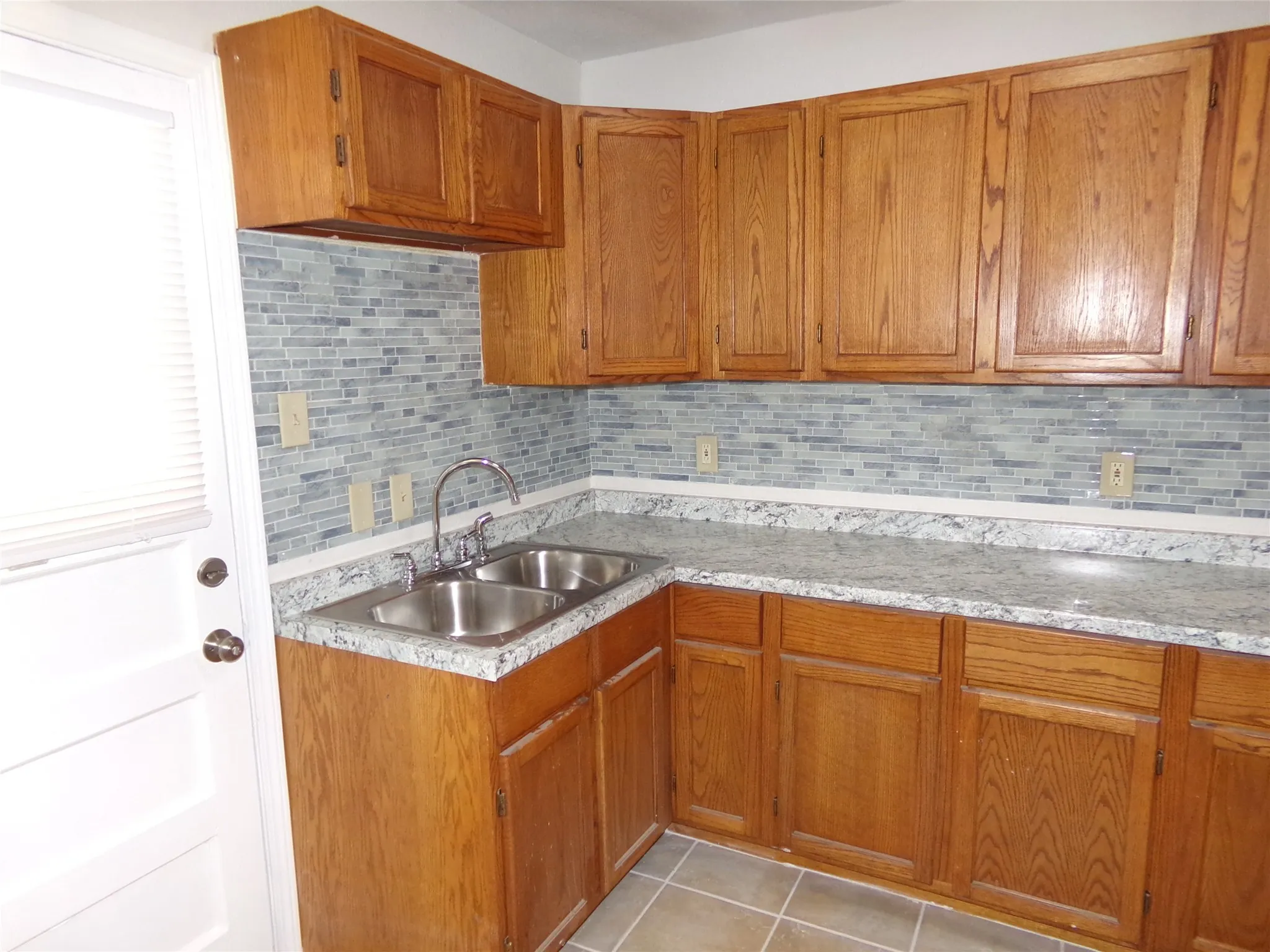Wood-finish cabinetry, mosaic tile backsplash, dual basin stainless steel sink, and light-toned countertops
