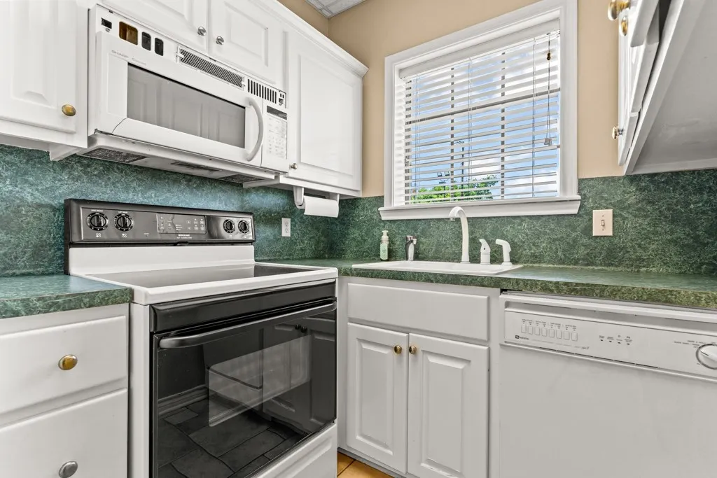 White cabinetry with gold-tone hardware, dark green countertops, white over-the-range microwave, built-in dishwasher, and white sink