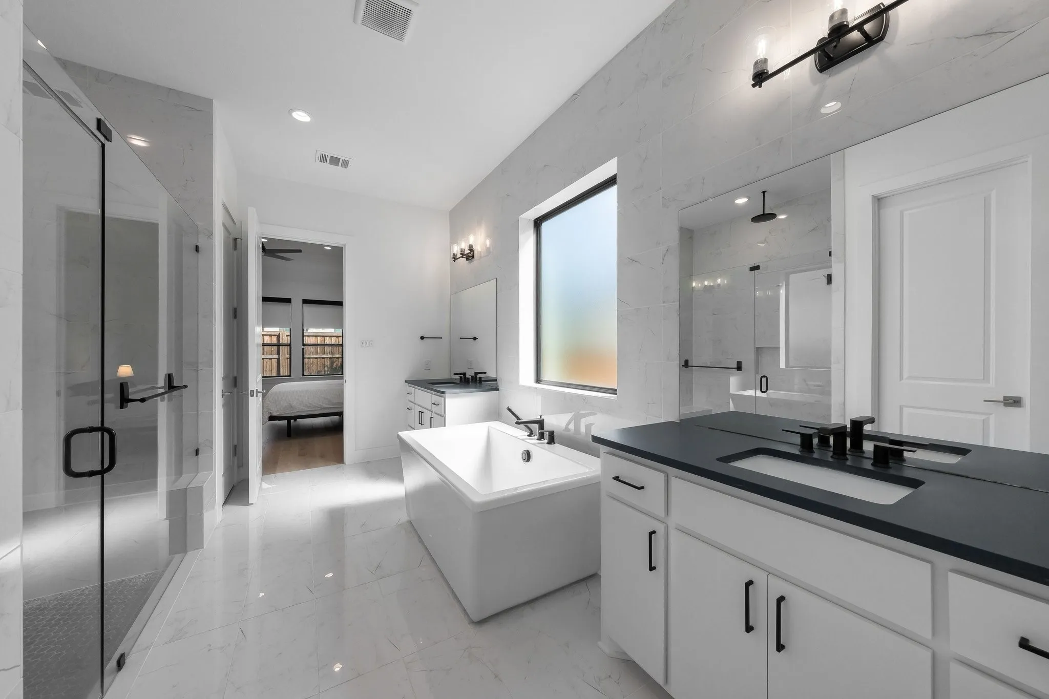 Spacious bathroom featuring white tiled flooring, a large freestanding soaking tub, a glass-enclosed shower, a double vanity with dark countertops, and a rectangular window