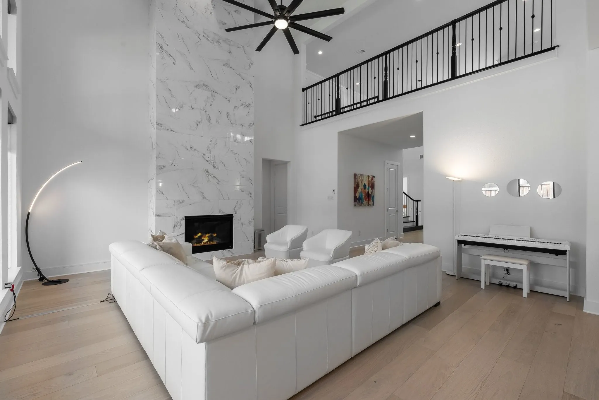 Vaulted ceiling with black ceiling fan, two-story fireplace with white marble-patterned surround, light wood-finish flooring, and black metal open-rail balcony