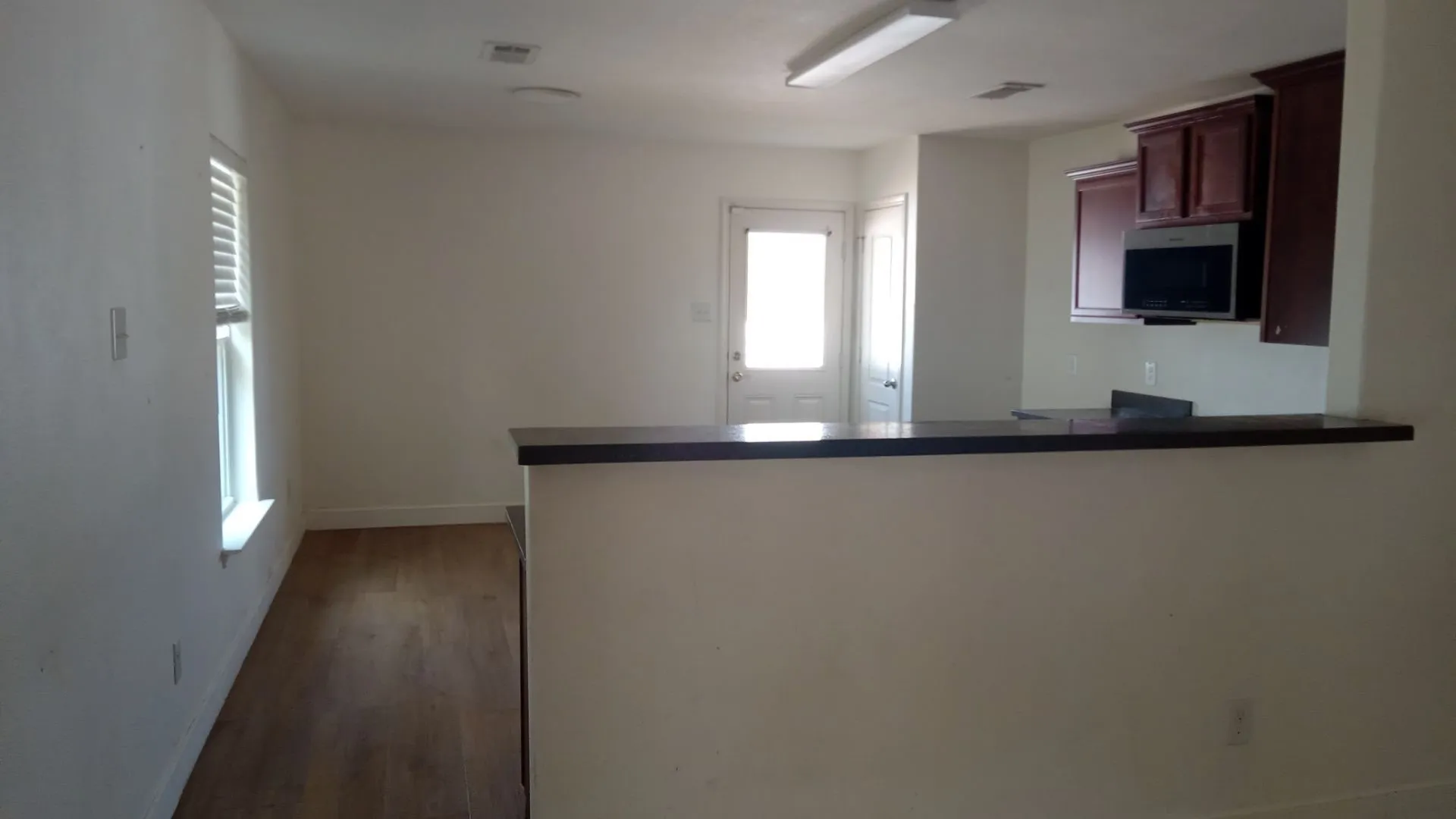 Wood-finish flooring, white window blinds, dark wood-finish cabinetry, stainless steel microwave, and a breakfast bar with a dark countertop