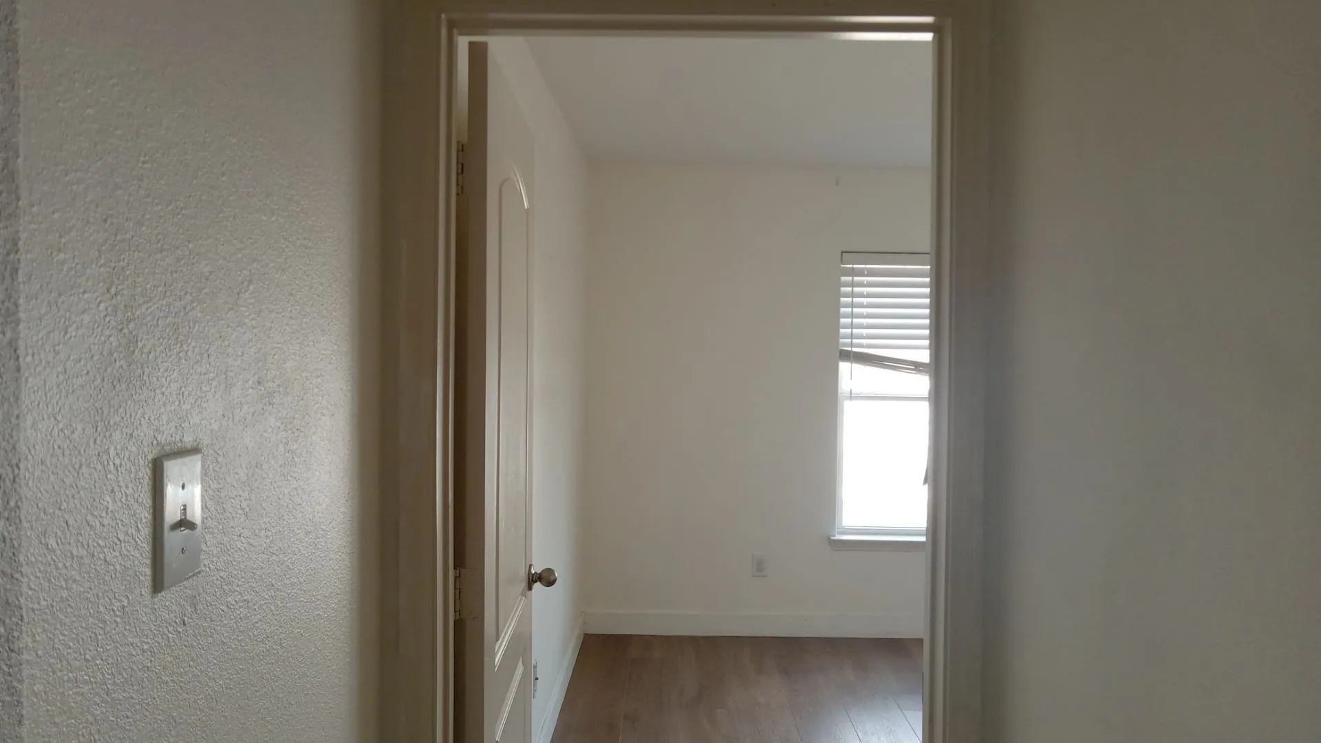 Room with wood-finish flooring, a window with horizontal blinds, and a white interior door