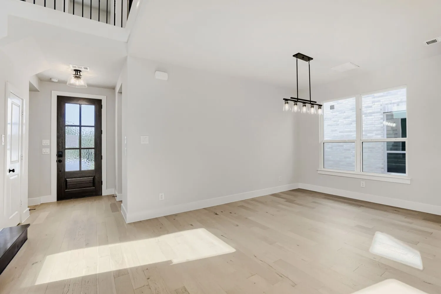 Wood-finish flooring throughout, wrought iron baluster staircase, dark wood entry door with frosted glass panes, modern linear chandelier, and multiple pane windows