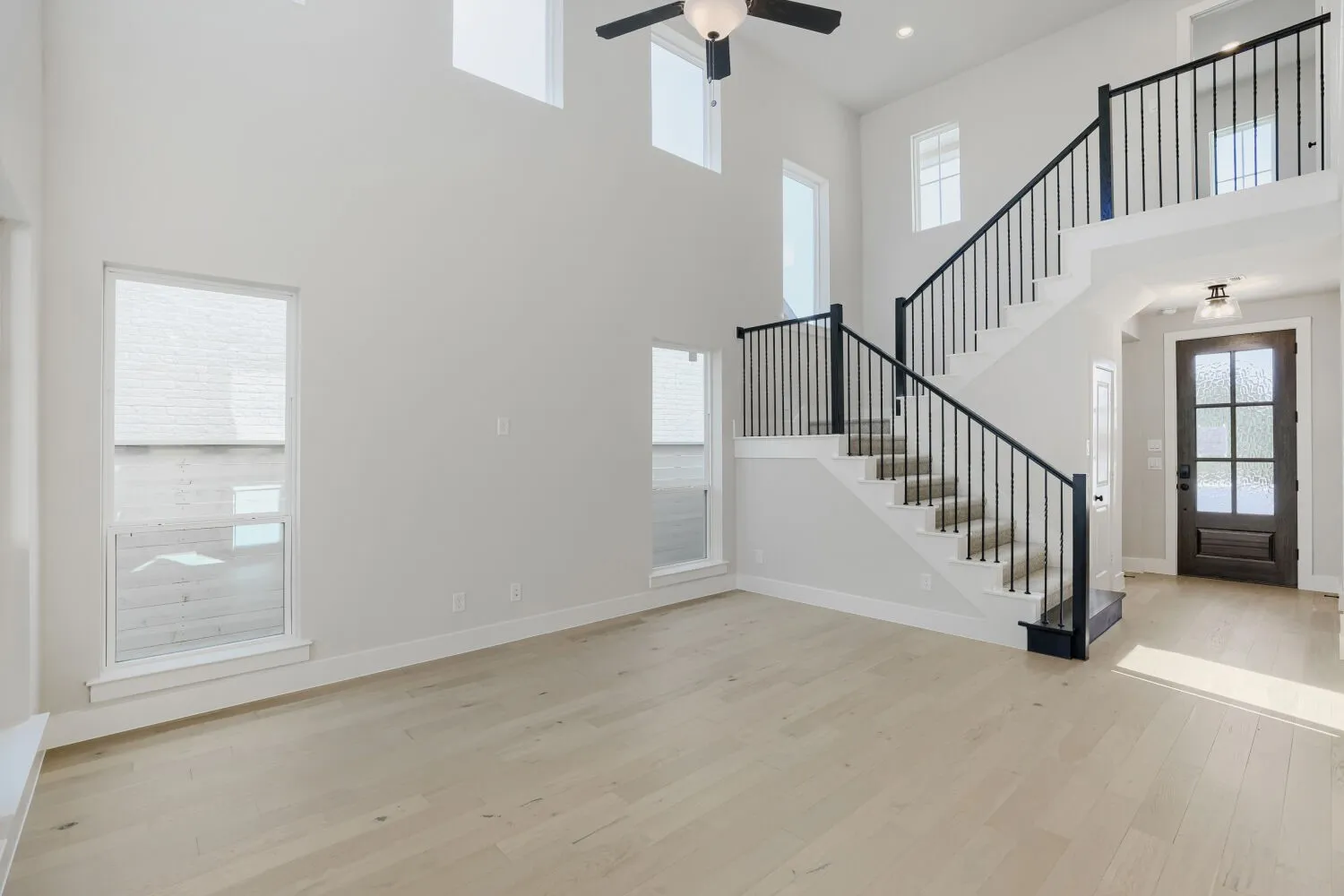 Double-height living space featuring light wood-finish flooring, multiple windows, a ceiling fan, and a grand staircase with dark iron balusters