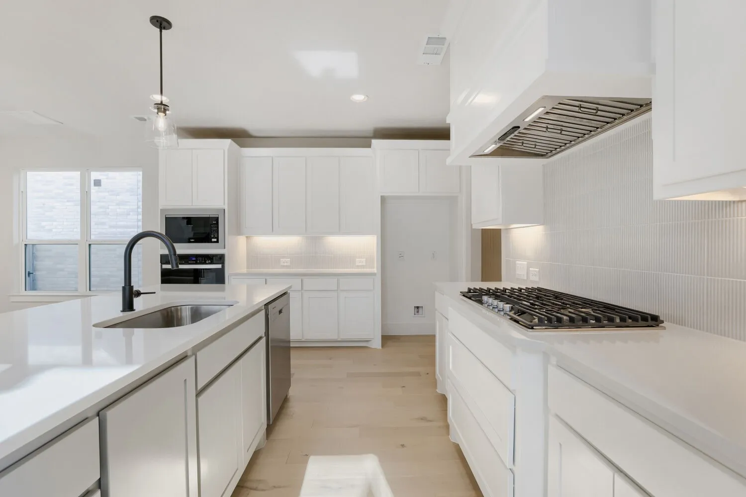 White cabinetry with under-cabinet lighting, light-toned wood-finish flooring, built-in microwave and oven, gas cooktop with custom range hood, and a kitchen island with an undermount sink and matte black faucet