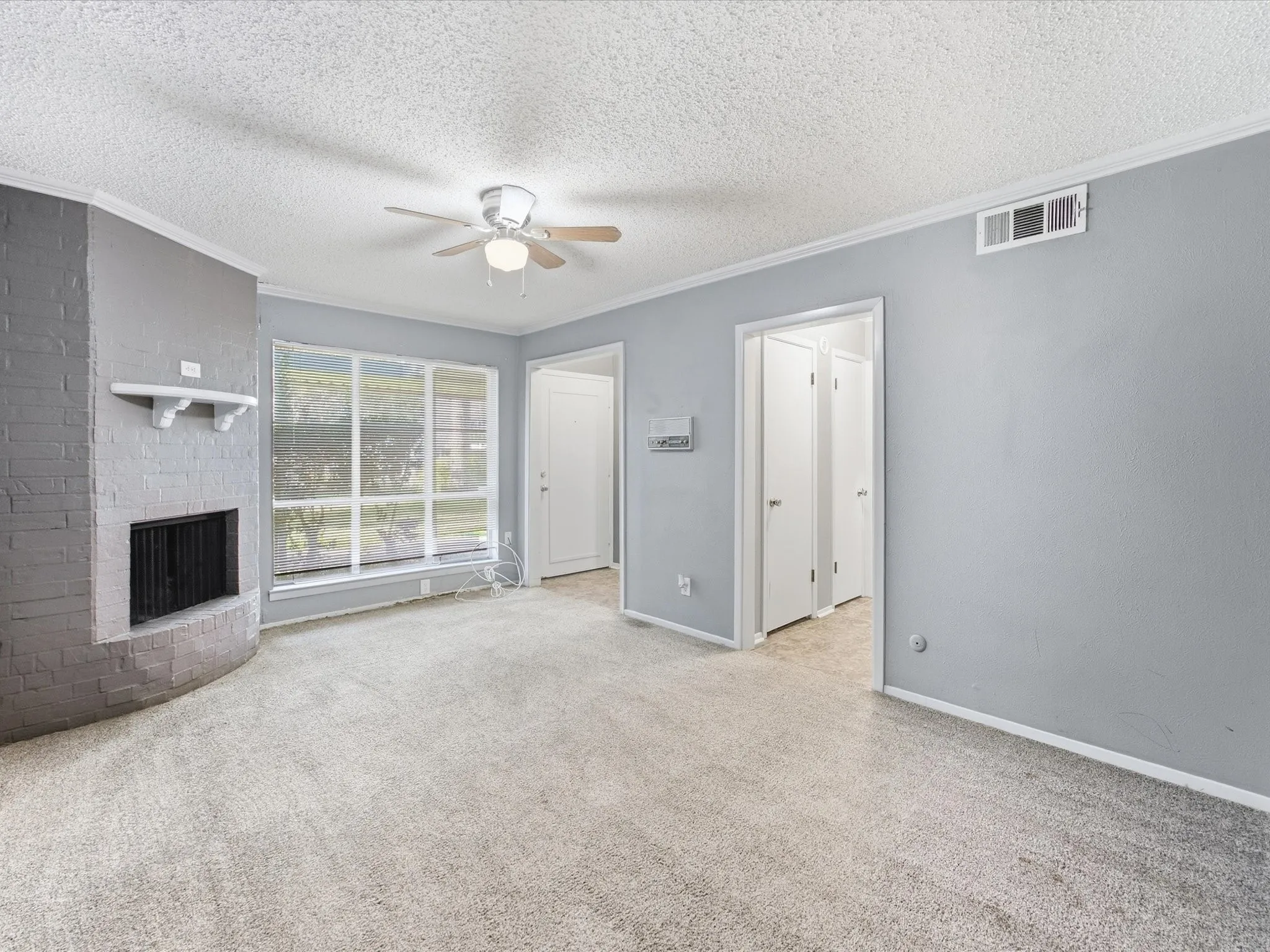 Corner brick fireplace with mantel, ceiling fan with integrated lighting, large window, and light-toned carpet