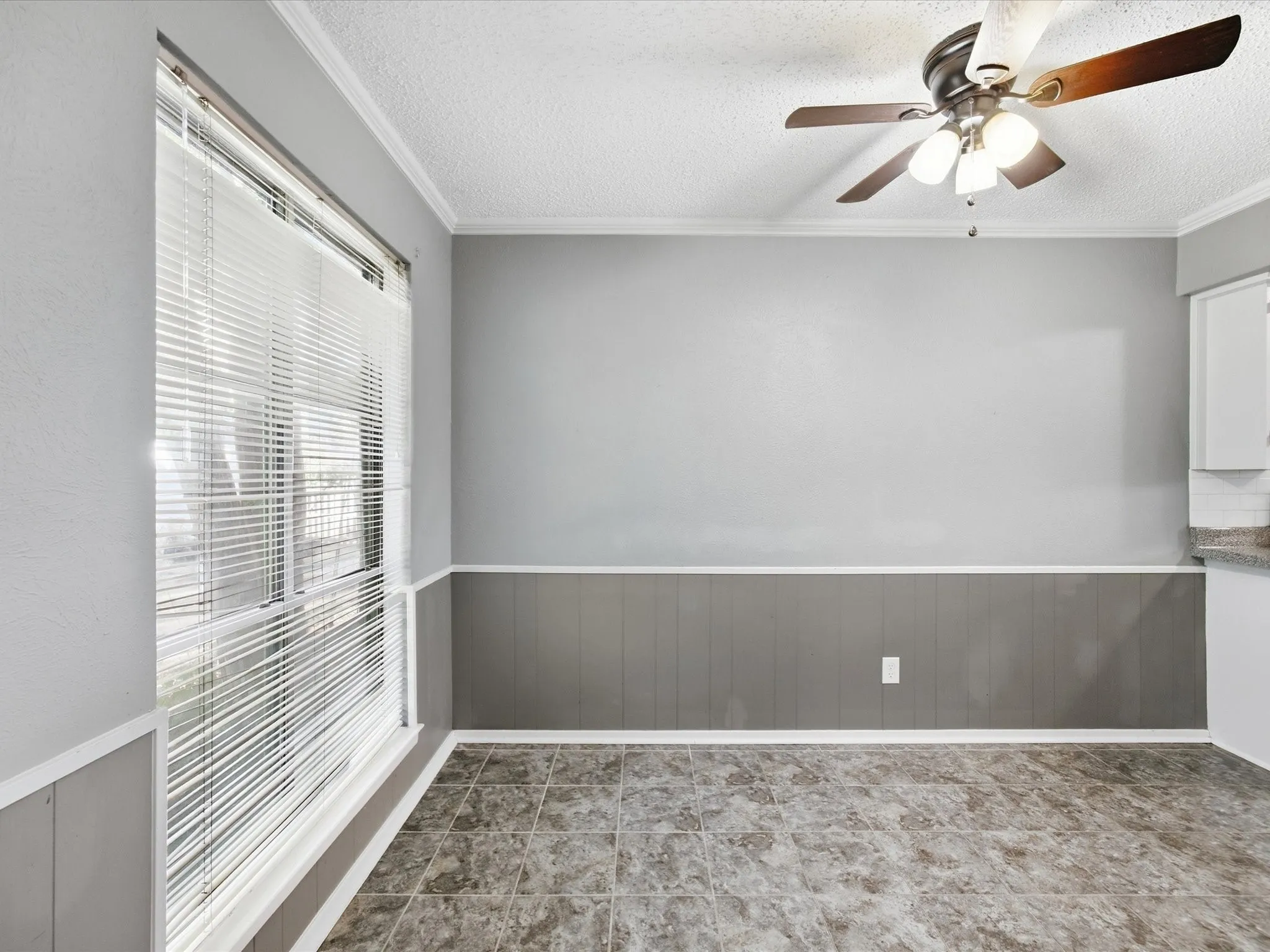 Recessed ceiling with crown molding, patterned tile flooring, ceiling fan with integrated lighting, and a large window with blinds