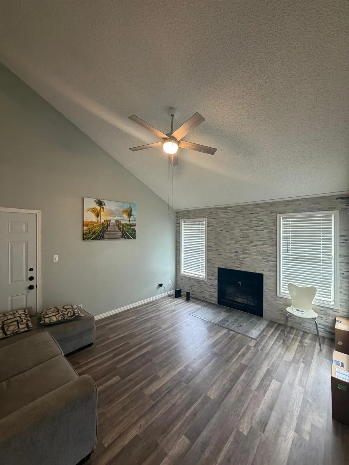 Vaulted ceiling with a ceiling fan, wood-finish flooring, fireplace with stone surround, and two windows with blinds