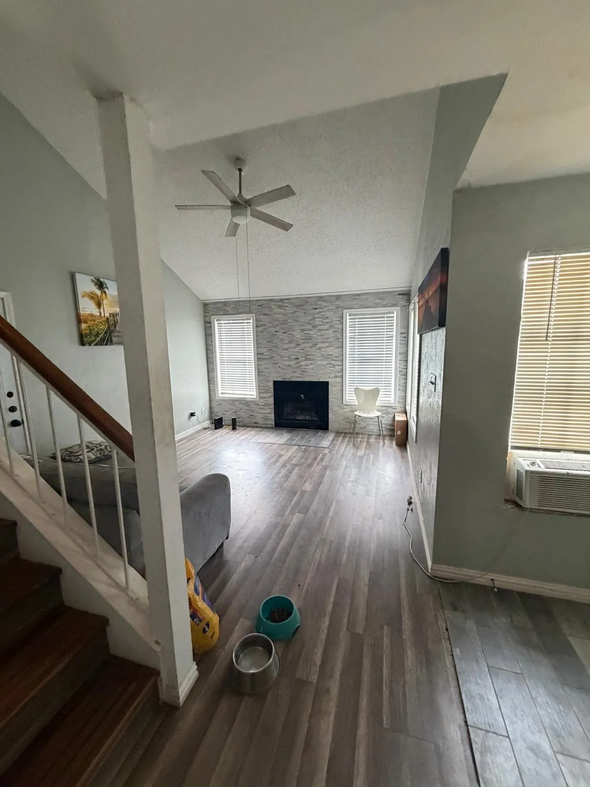 Vaulted ceiling, wood-finish flooring, fireplace with a stone-finish surround, two windows with blinds, and a ceiling fan