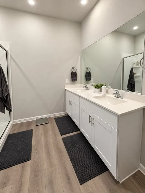 Double vanity with white cabinetry and integrated sinks, extensive wall-mounted mirror, recessed lighting, and wood-finish flooring