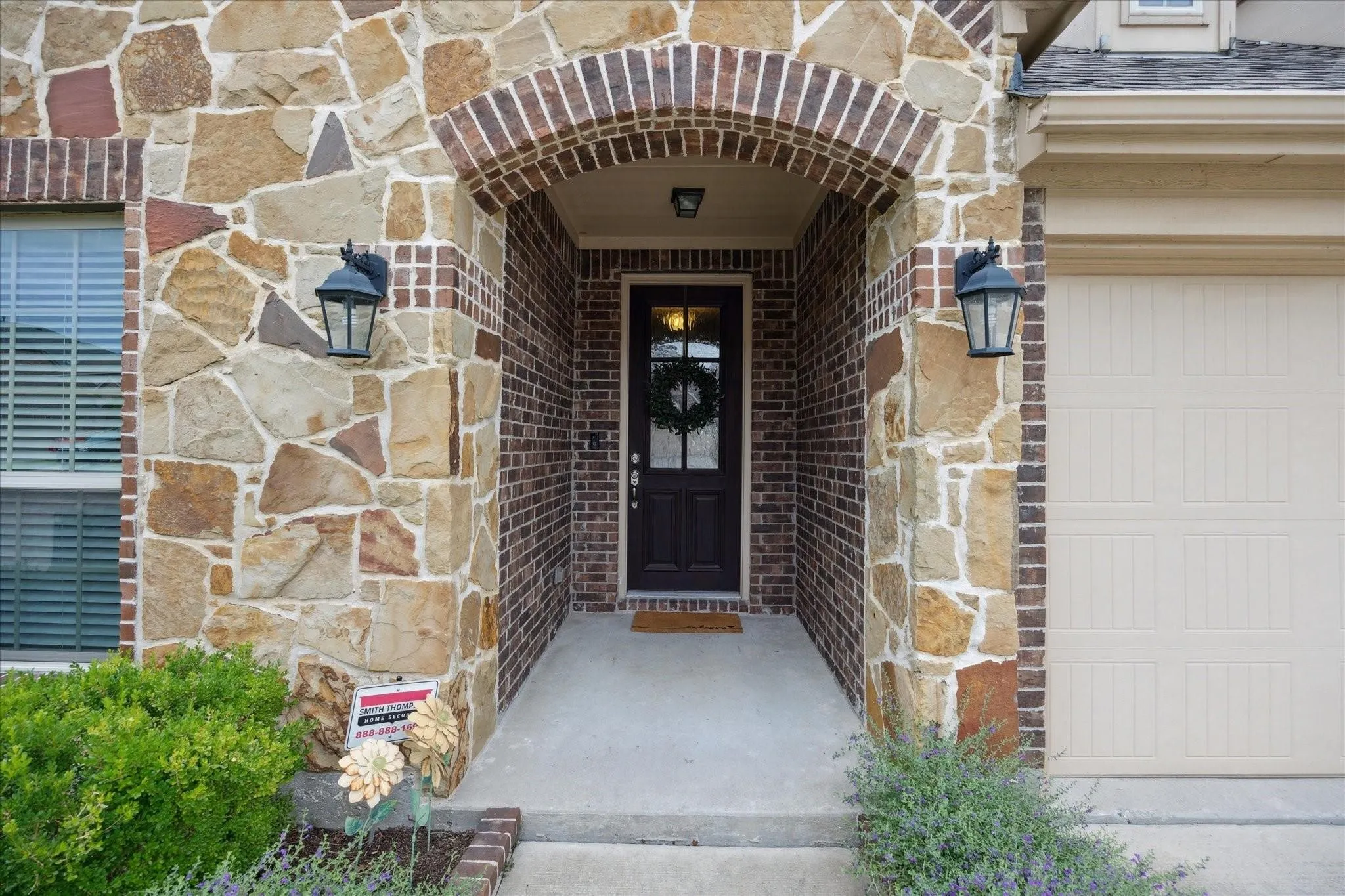 Stone and brick facade with an arched entryway, featuring a dark panel door with glass inserts, exterior sconces, and an attached garage