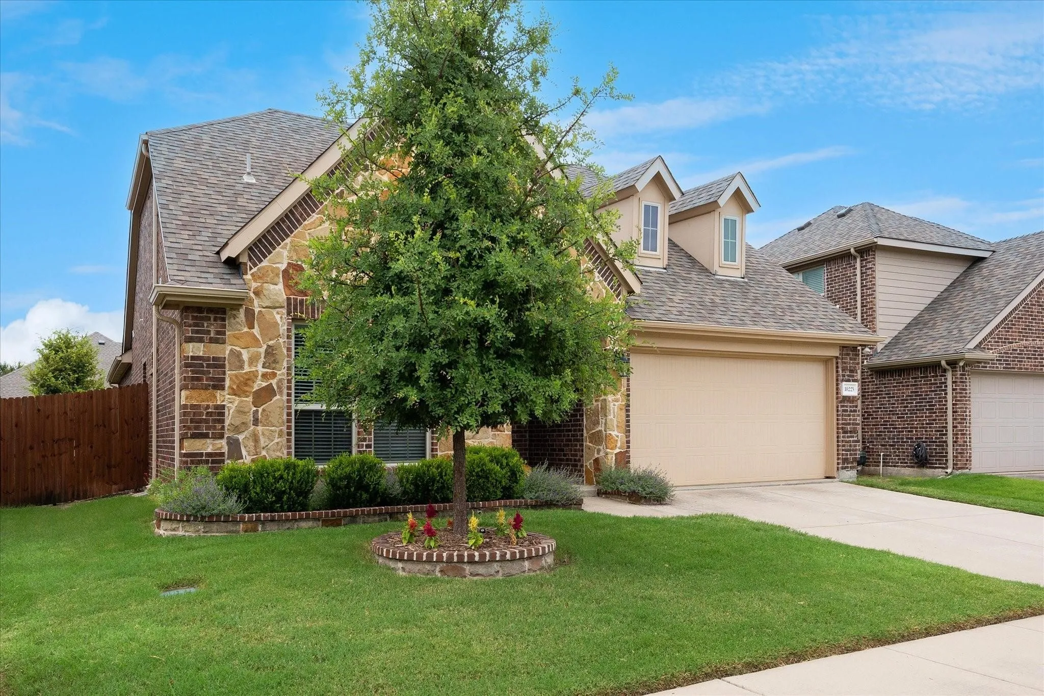 Brick and stone exterior with a two-car garage, dormer windows, and a manicured lawn featuring a planted tree