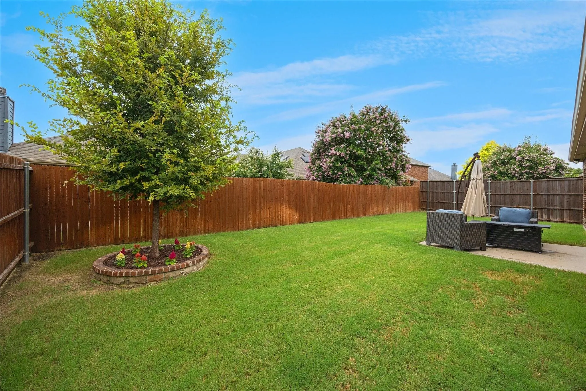 Expansive backyard featuring a manicured lawn, established tree with brick-lined flower bed, and a concrete patio