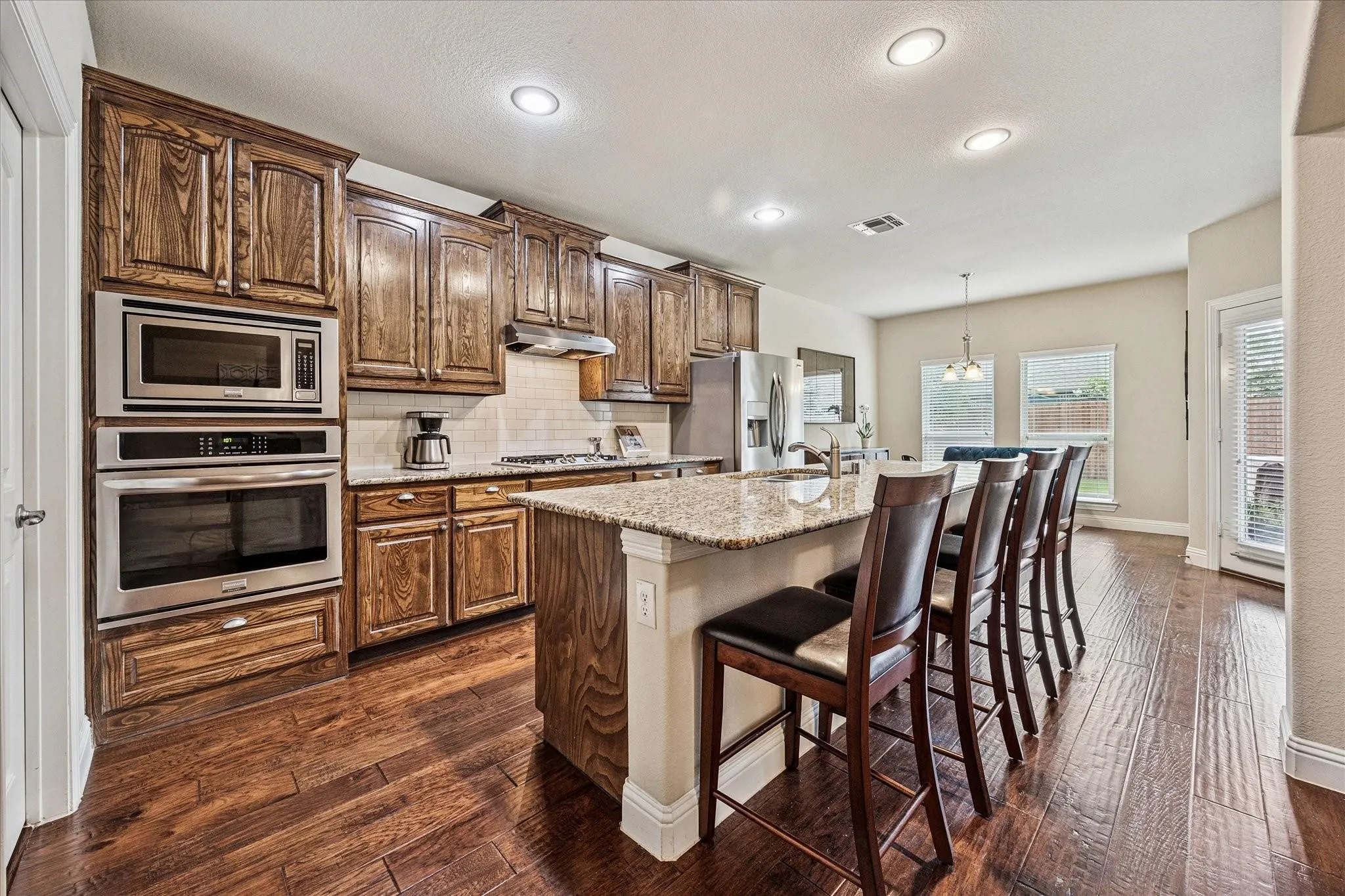 Kitchen featuring wood-finish flooring, built-in stainless steel appliances, stained wood cabinetry, a tiled backsplash, and a granite-finish island with seating