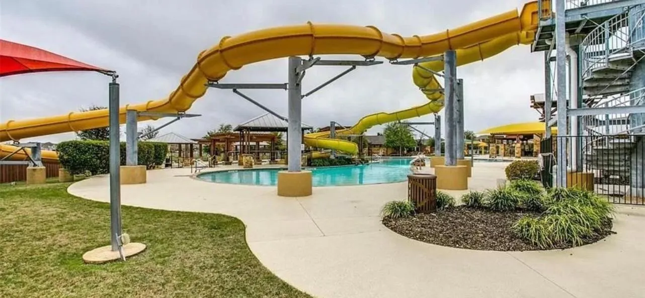 Swimming pool with curved edges, multiple yellow waterslides, expansive concrete decking, integrated landscaping, and shaded pergolas