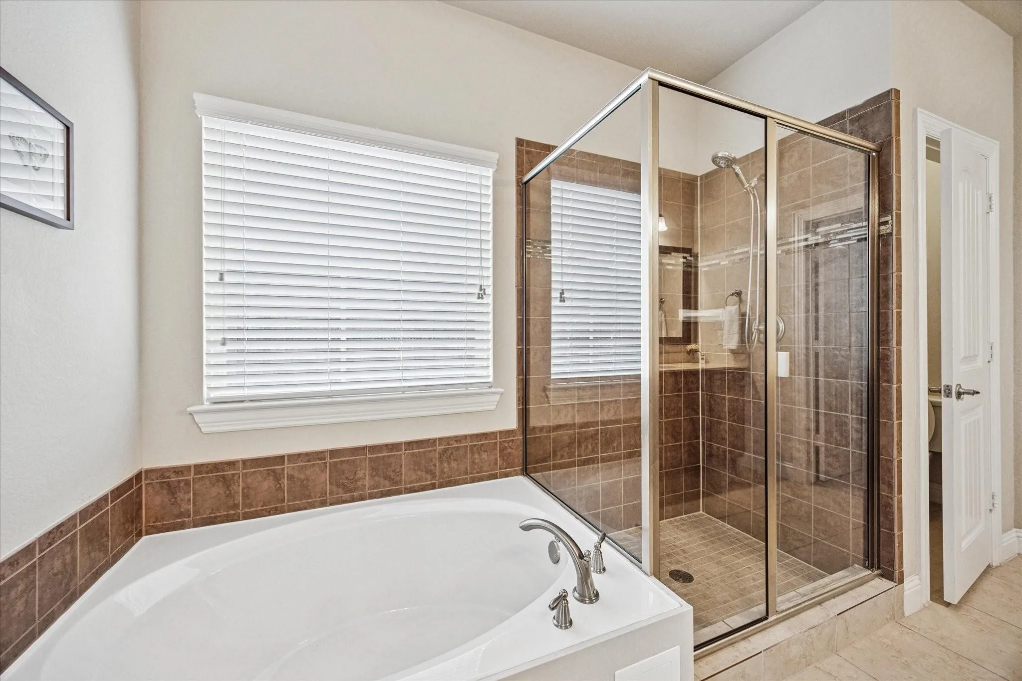 White soaking tub with brushed nickel fixtures, glass-enclosed shower with tiled surround, and a window with white blinds