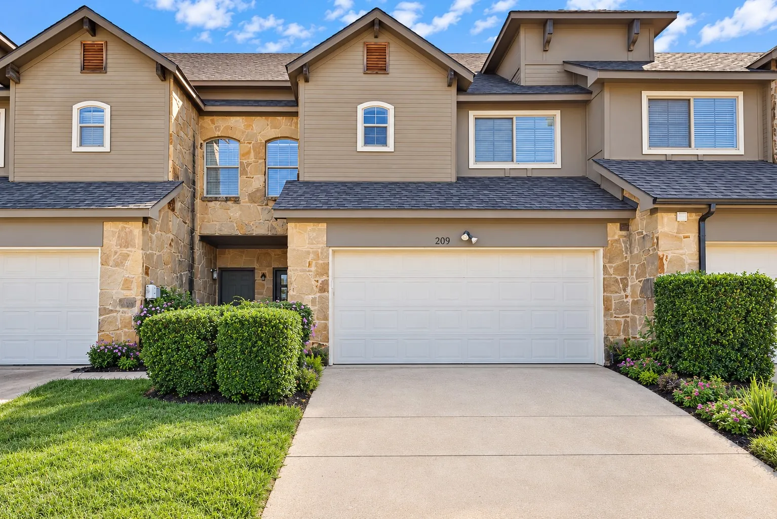 Front exterior: Two-story townhome with stone and siding facade and a two-car attached garage. Manicured lawn and landscaped beds.