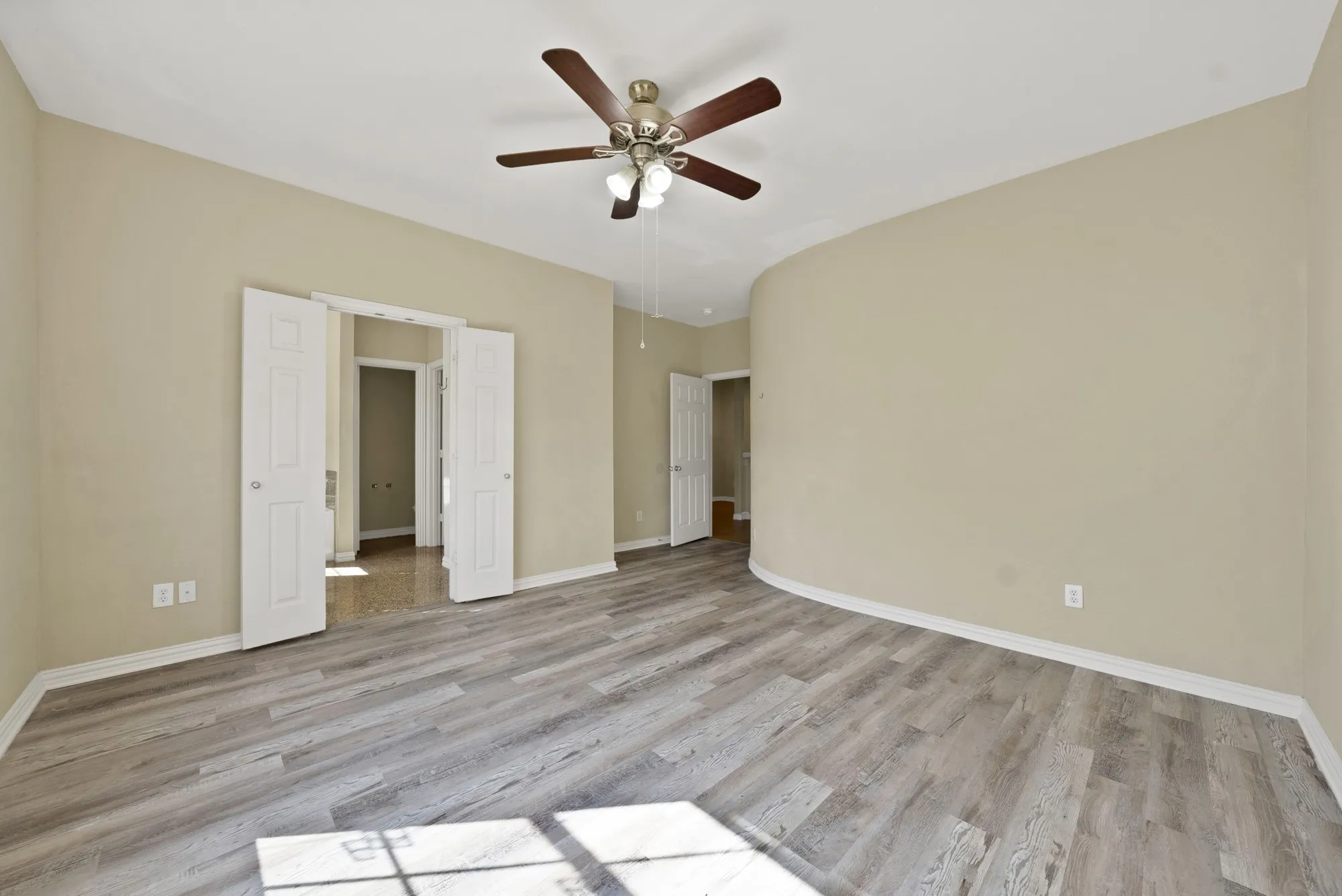 Upstairs primary bedroom with ensuite bathroom Ceiling fan, wide window with blinds, and wood-look flooring. Double doors on the right side of the room.