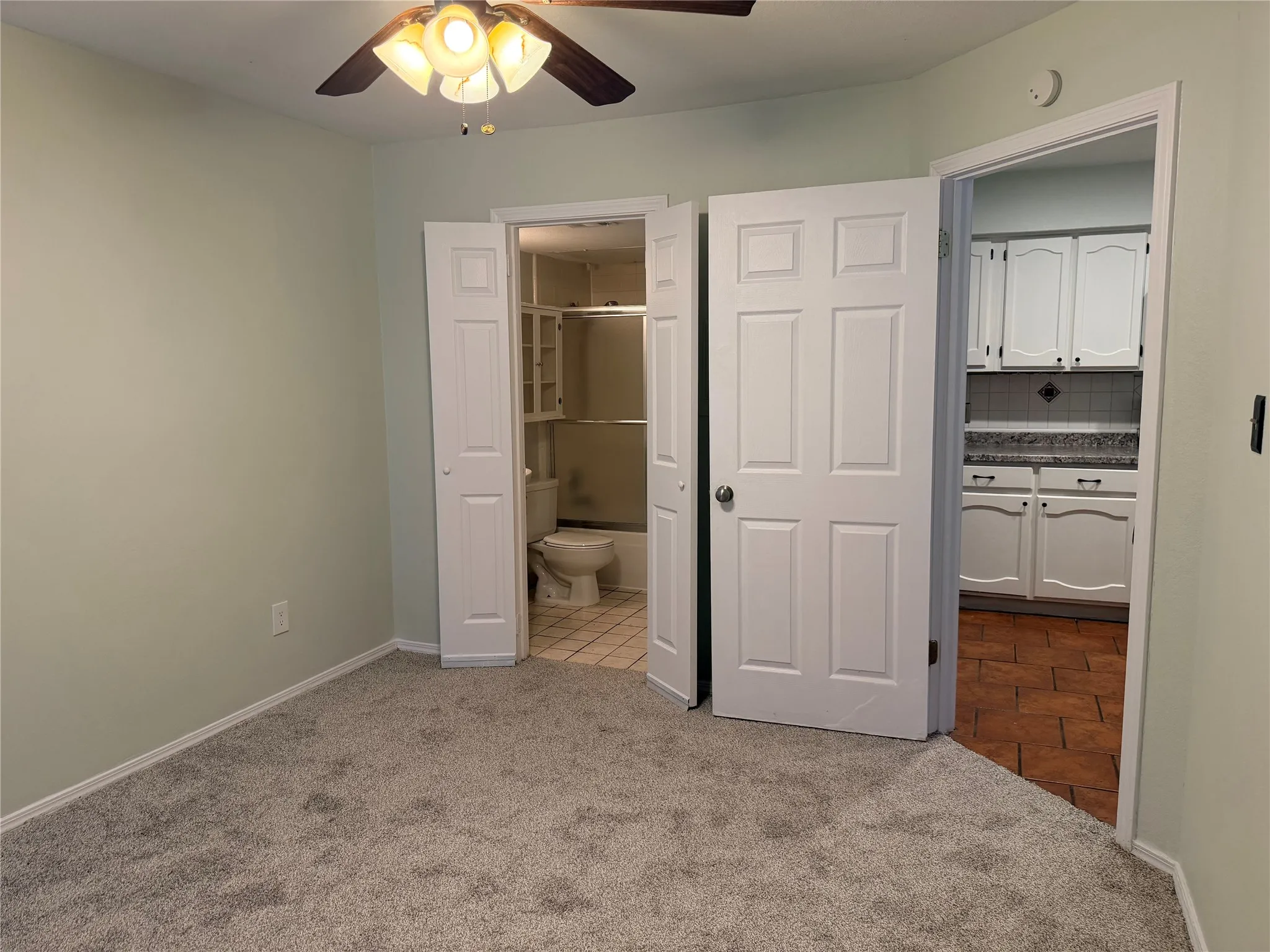 Plush carpet flooring, light-toned wall paint, ceiling fan, white paneled doors, and a bathroom with tile flooring