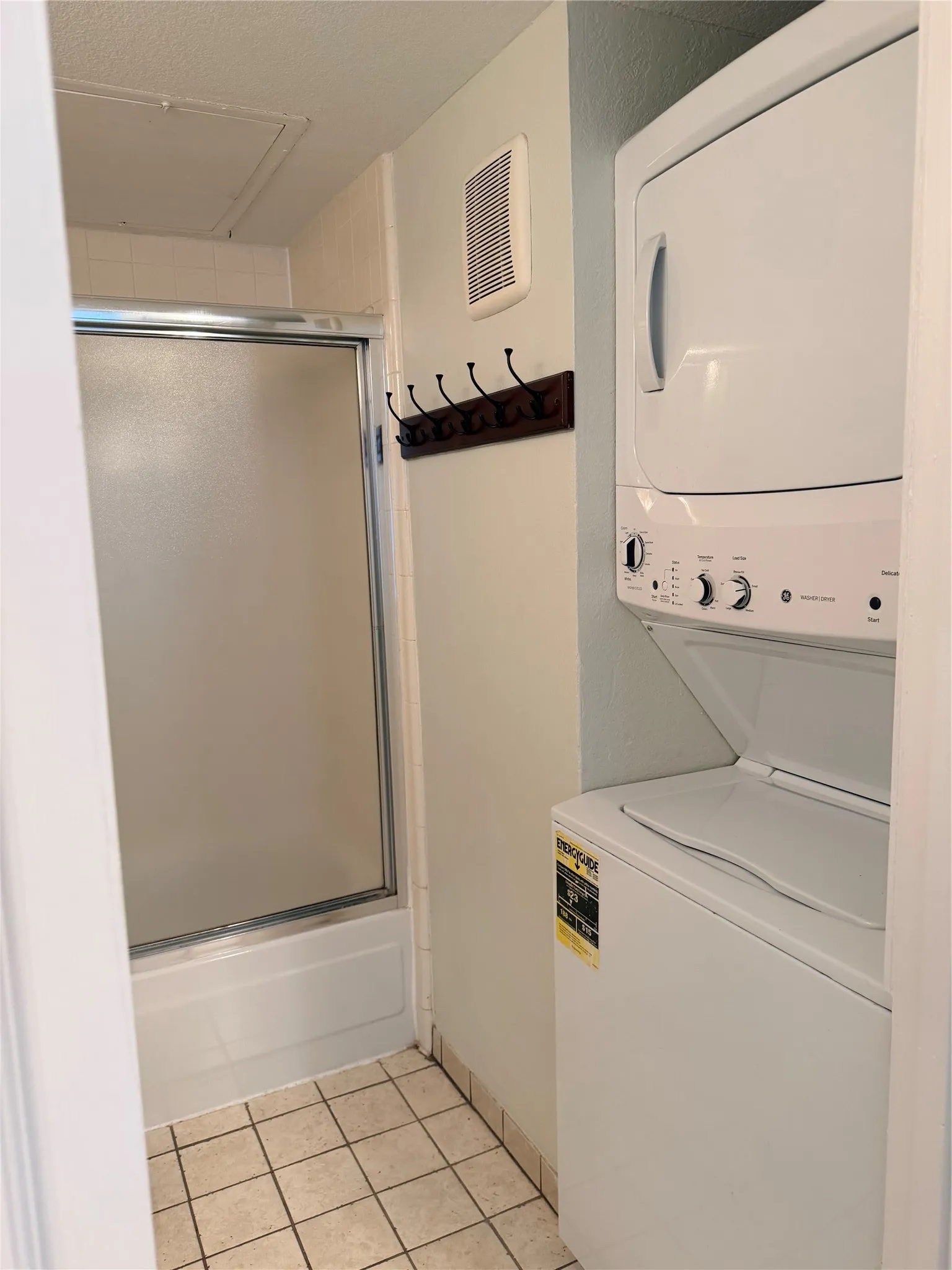 Stacked washer and dryer unit, tiled flooring, glass shower enclosure, and built-in wall vent