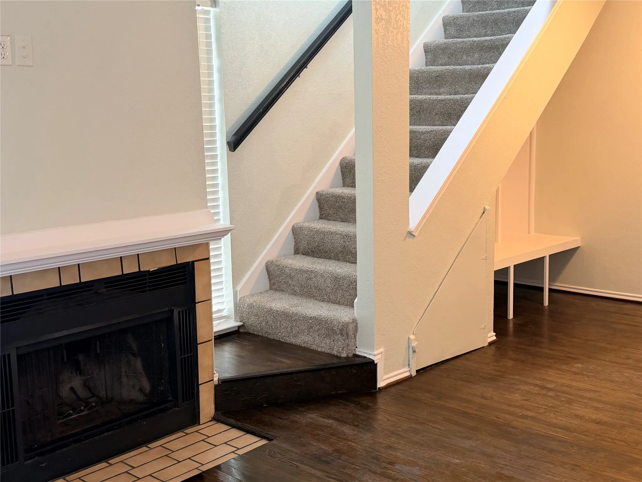 Wood-finish flooring, staircase with carpeted treads and dark handrail, fireplace with tile surround and mantel, and built-in under-stair bench