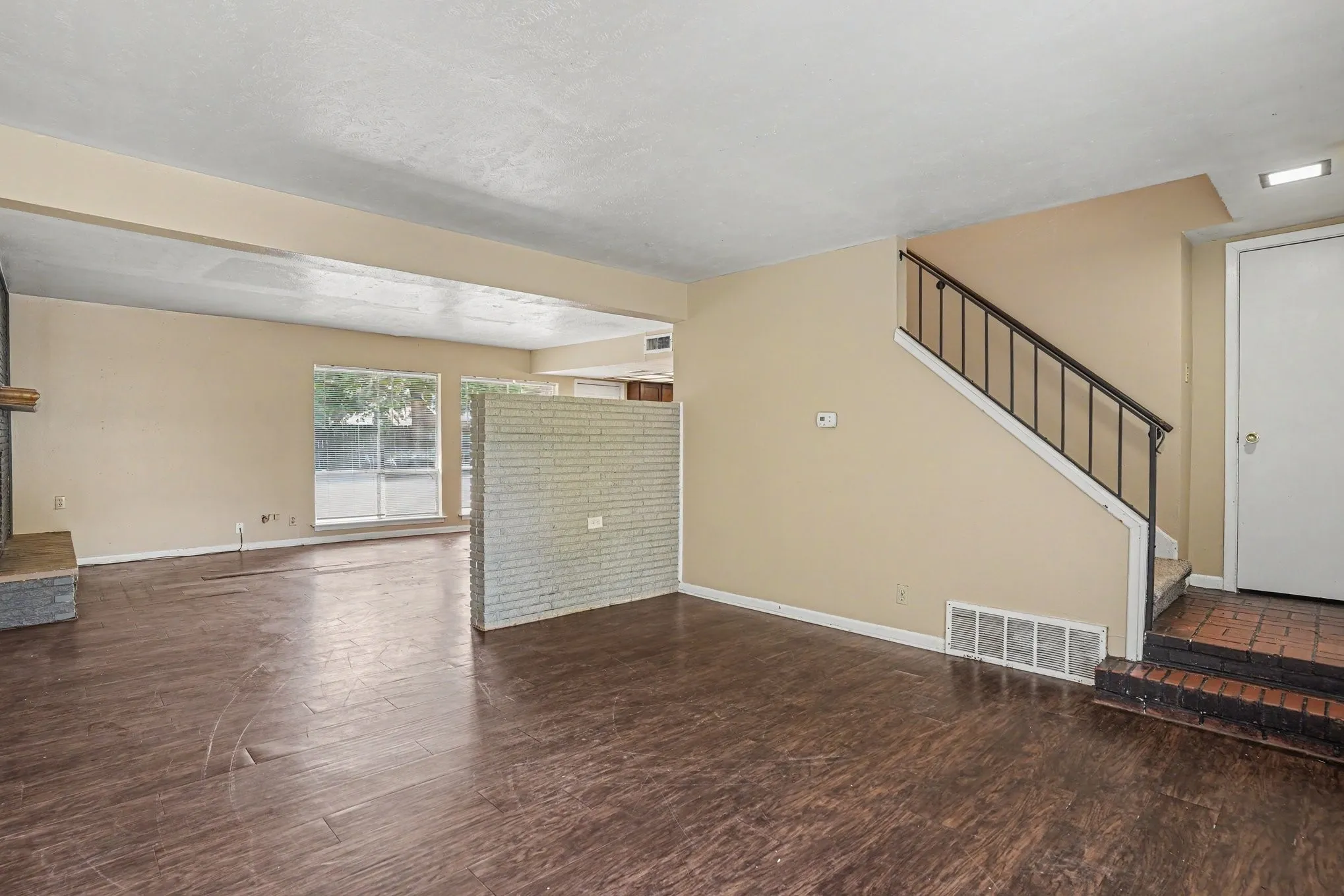 Wood-finish flooring throughout, brick accent partition, fireplace with stone surround, large window with outdoor visibility, and a staircase with wrought iron railing