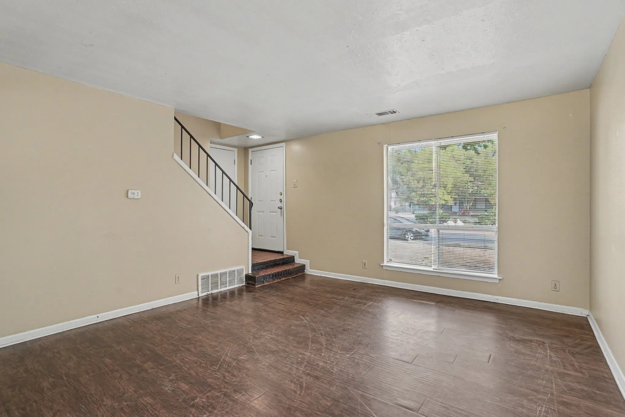 Spacious living area featuring wood-finish flooring, a large window, neutral wall tones, and a staircase with a black metal railing