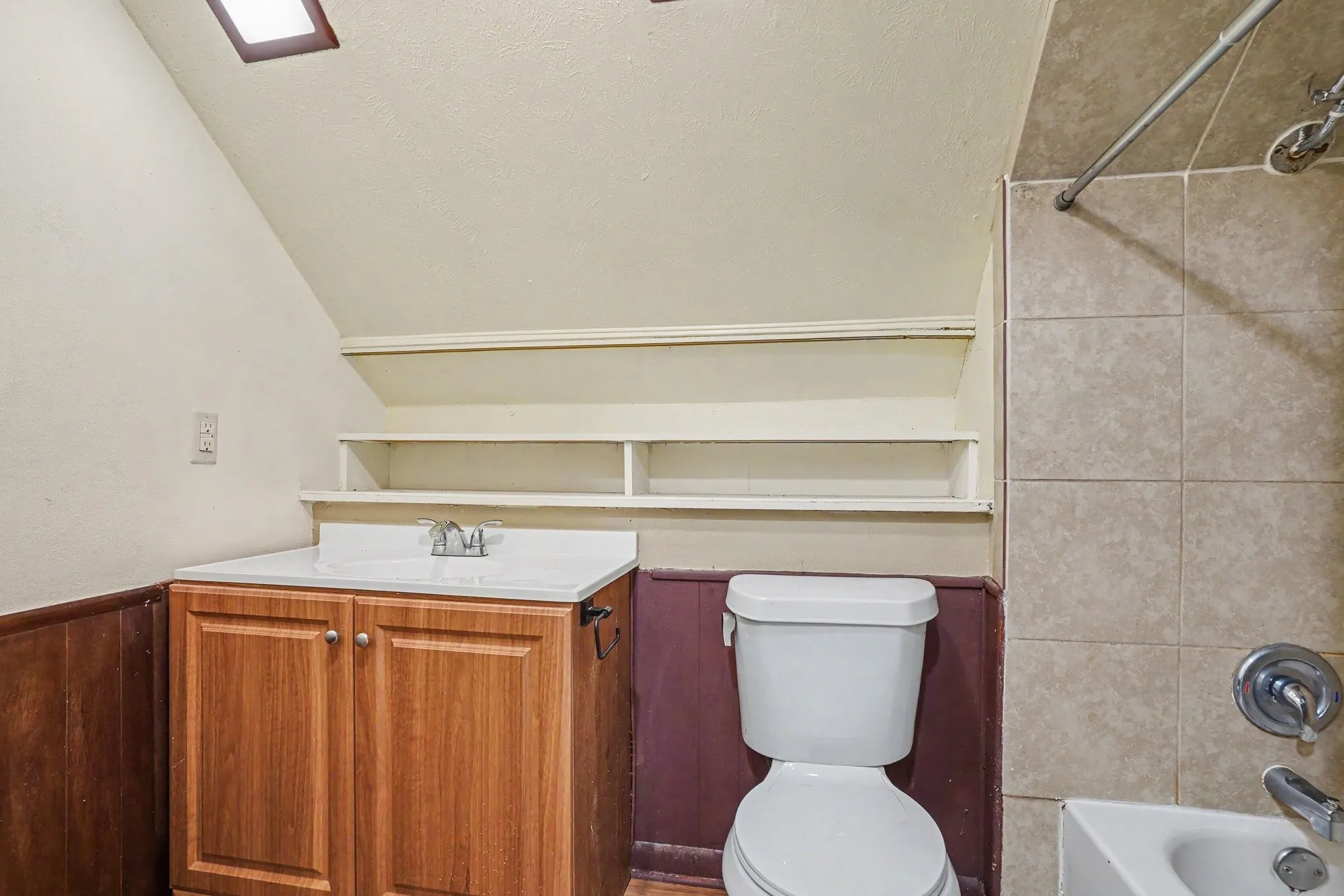 White vanity countertop, wood-finish vanity cabinetry, built-in shelving, tiled shower surround, and a skylight