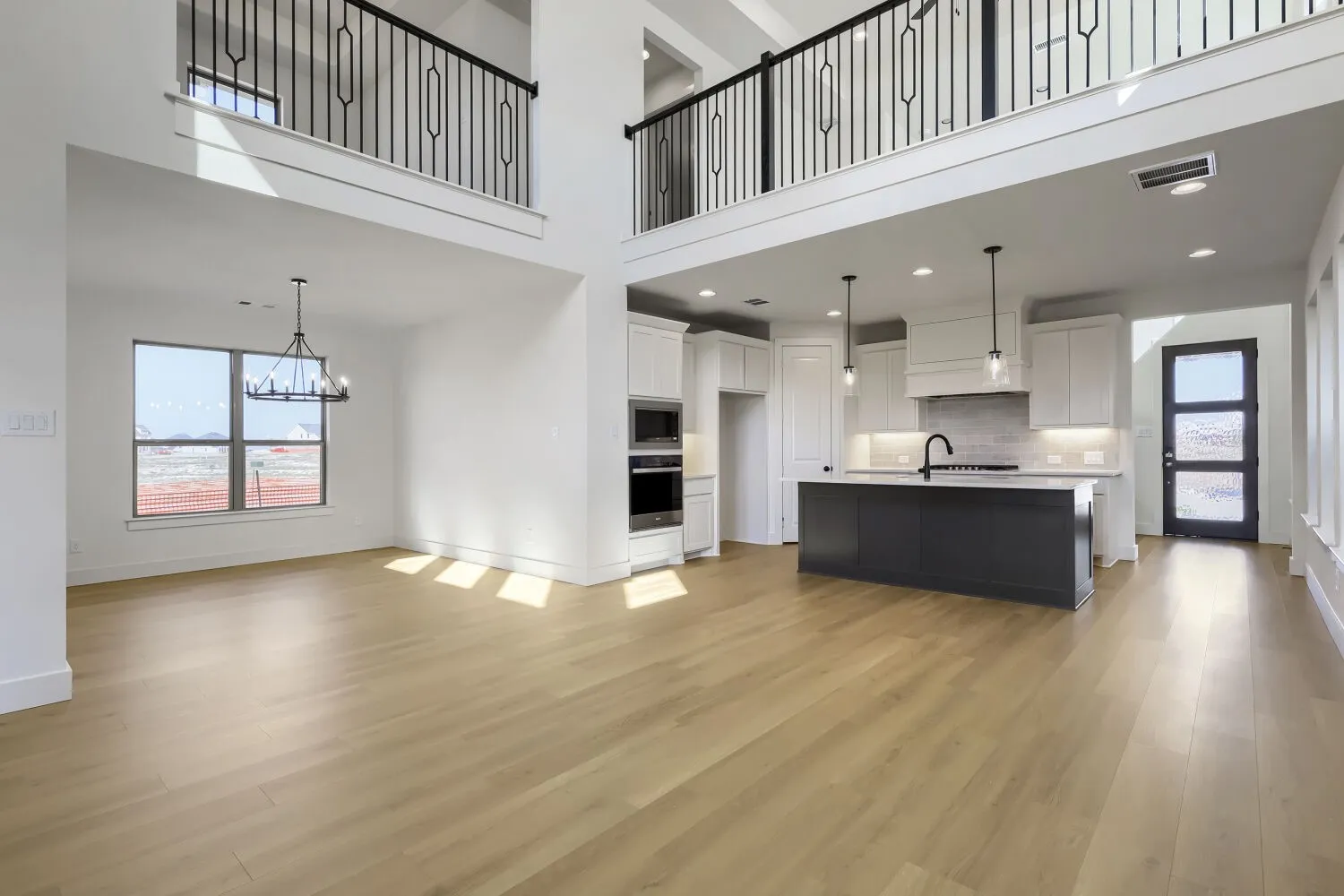 Open-concept living space featuring soaring two-story ceilings with mezzanine-level railings, abundant natural light from large windows, and wide-plank wood-finish flooring throughout