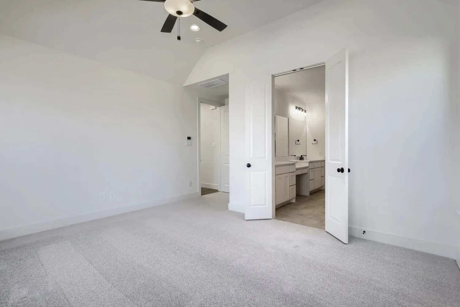 Vaulted ceiling with a flush-mount ceiling fan, light gray carpeting, and double panel doors leading to a bathroom with dual vanities and a tile floor