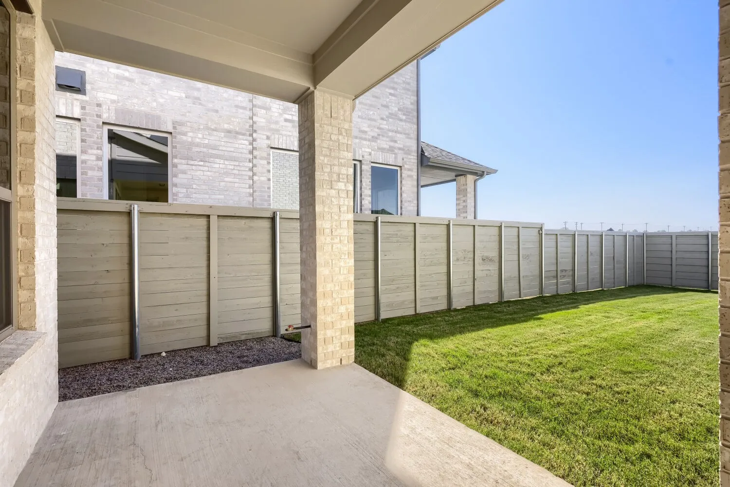Covered patio with concrete flooring, framed by brick columns, opening to a fenced yard with natural grass