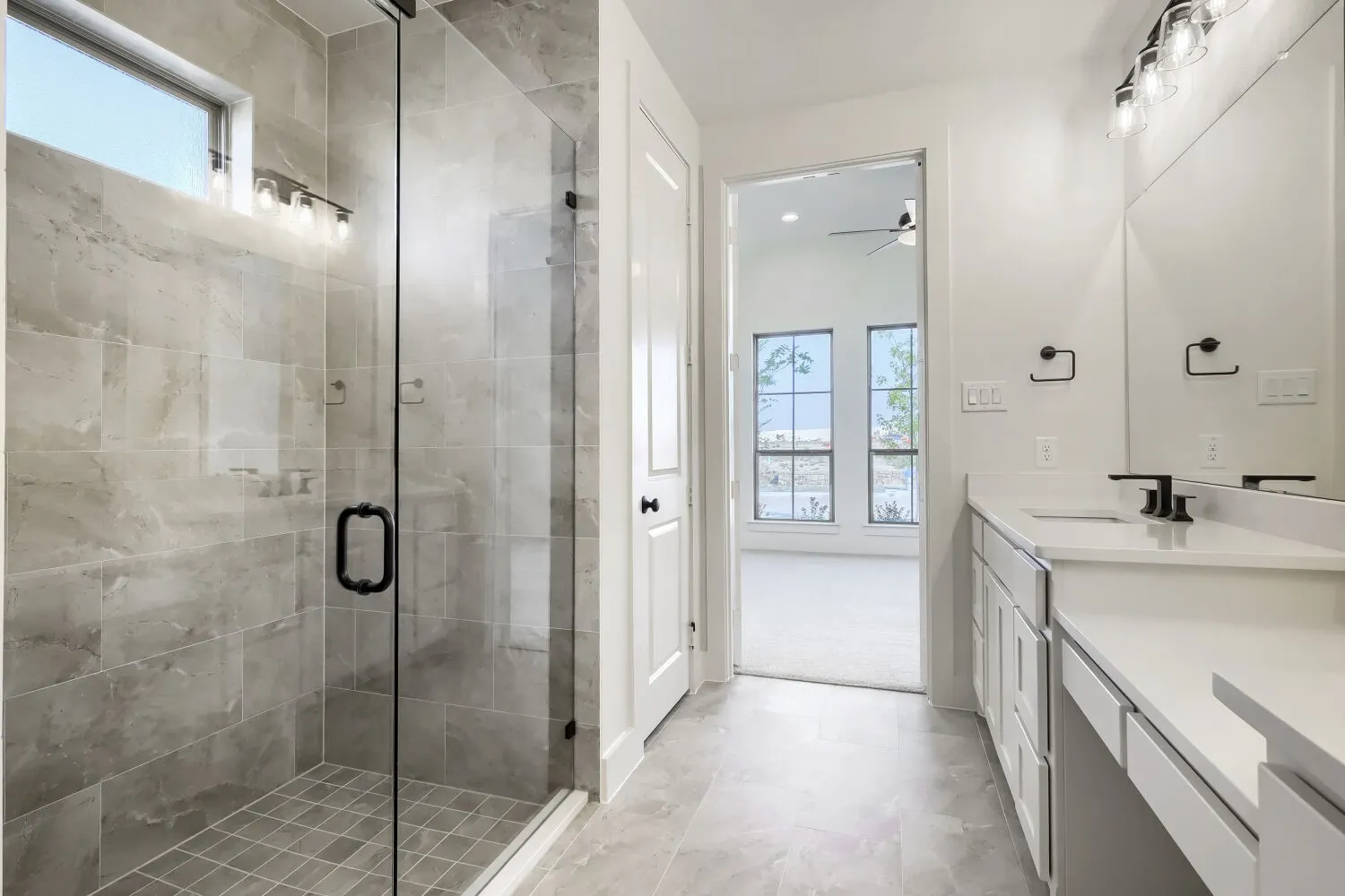 Spa-inspired primary bathroom featuring a frameless glass-enclosed walk-in shower with stone-finish tile surround, dual vanities with white cabinetry and matte black hardware, polished stone flooring, and abundant natural light from transom windows